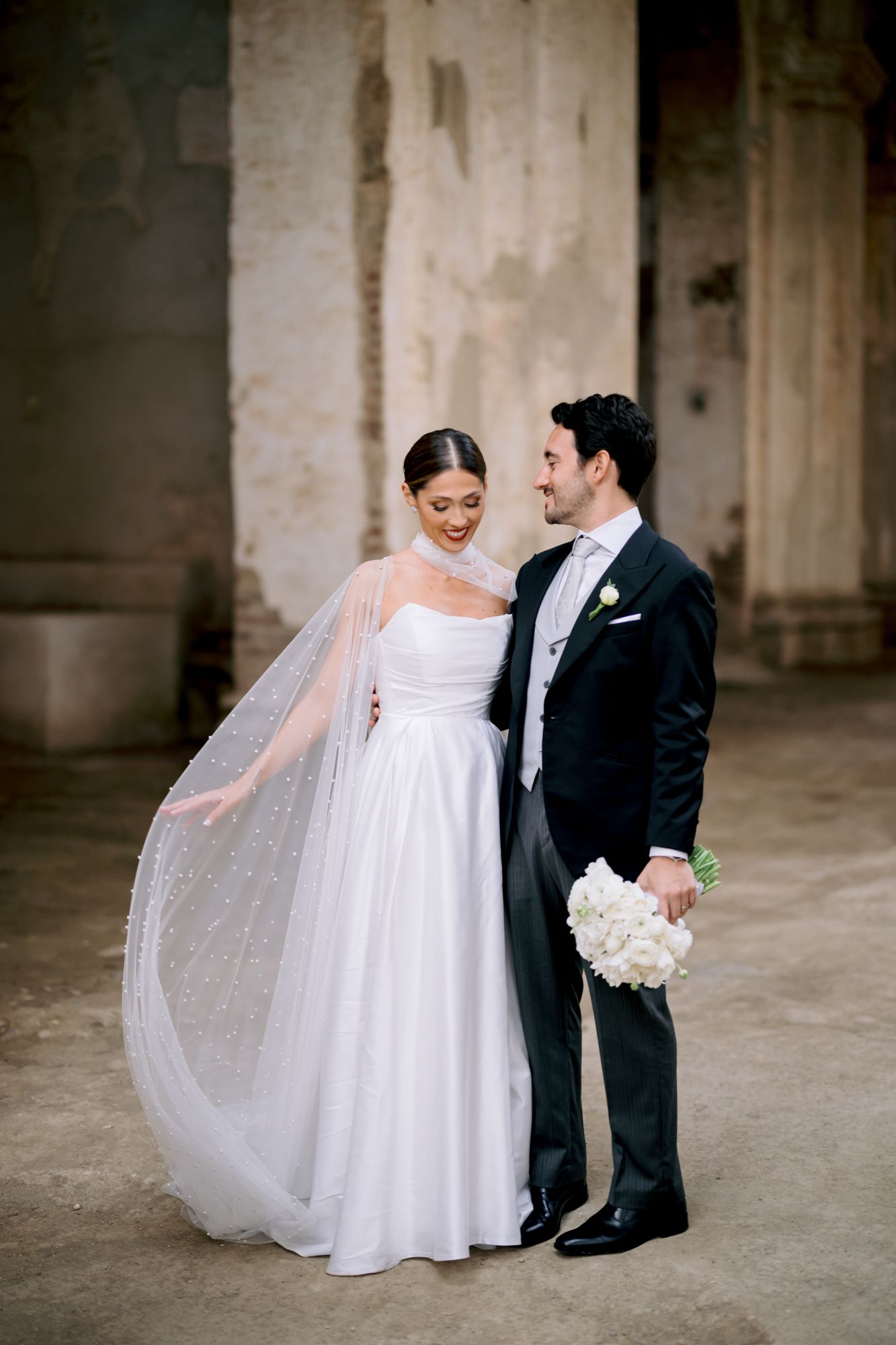Bride in classic white gown with sparkly cape stands close to groom holding white flowers.