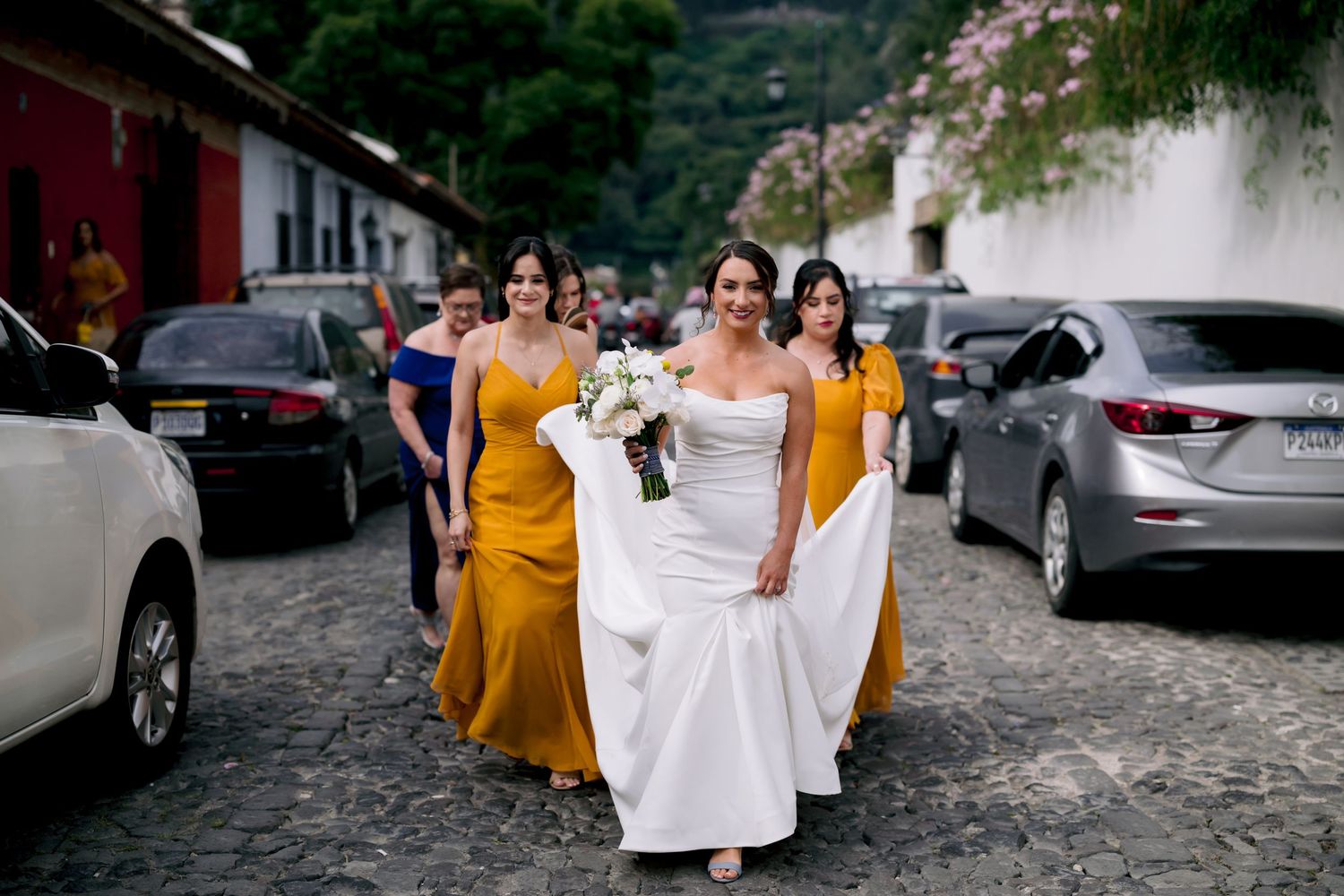 Bride in white gown walking with bridesmaids in yellow dresses along a cobblestone street.