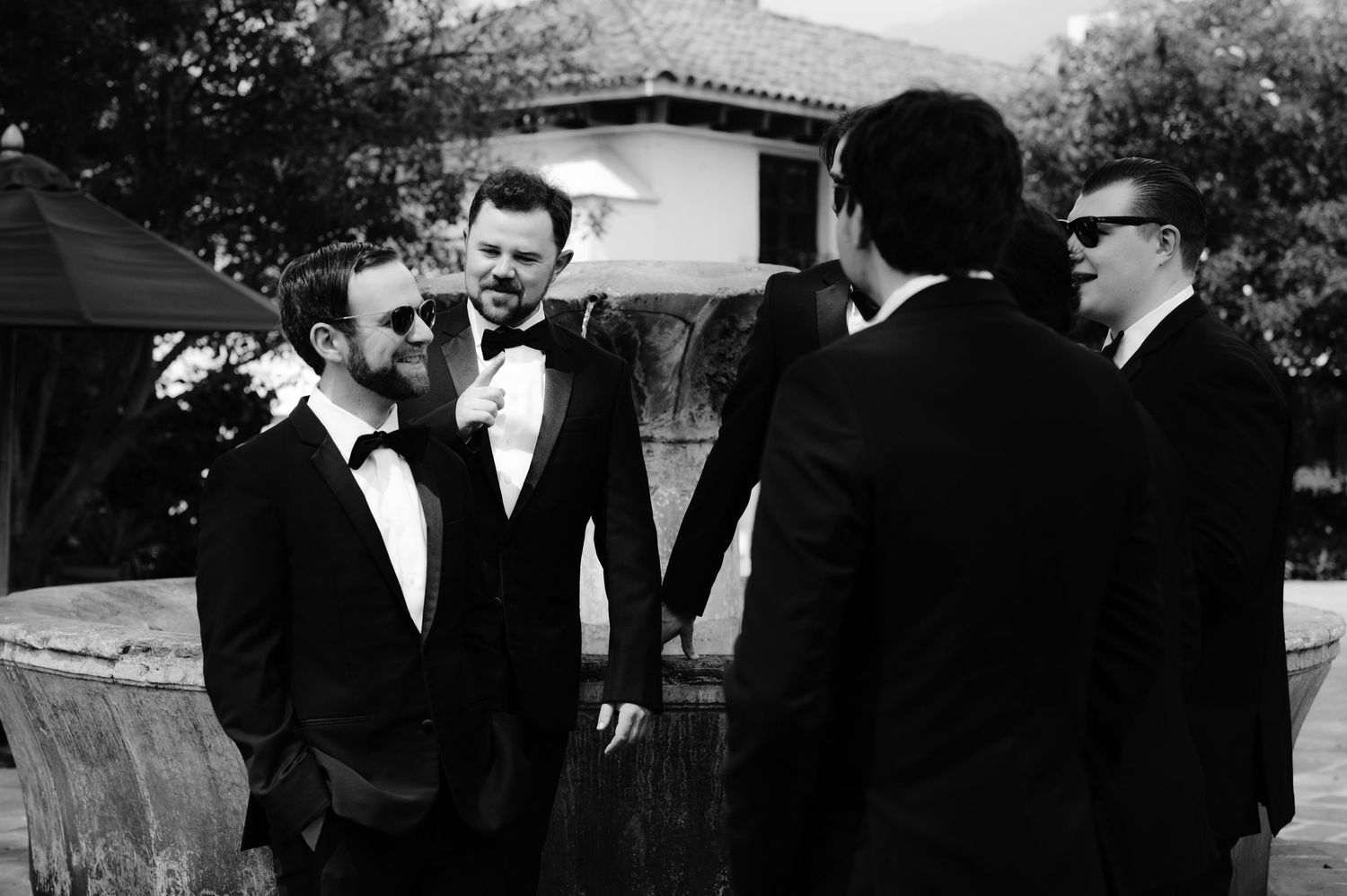 Black and white photo of groomsmen in tuxedos sharing a casual moment outdoors.