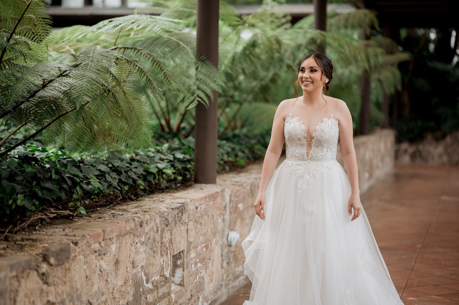 Elegant bride in white tulle gown stands on stone pathway surrounded by tropical plants.