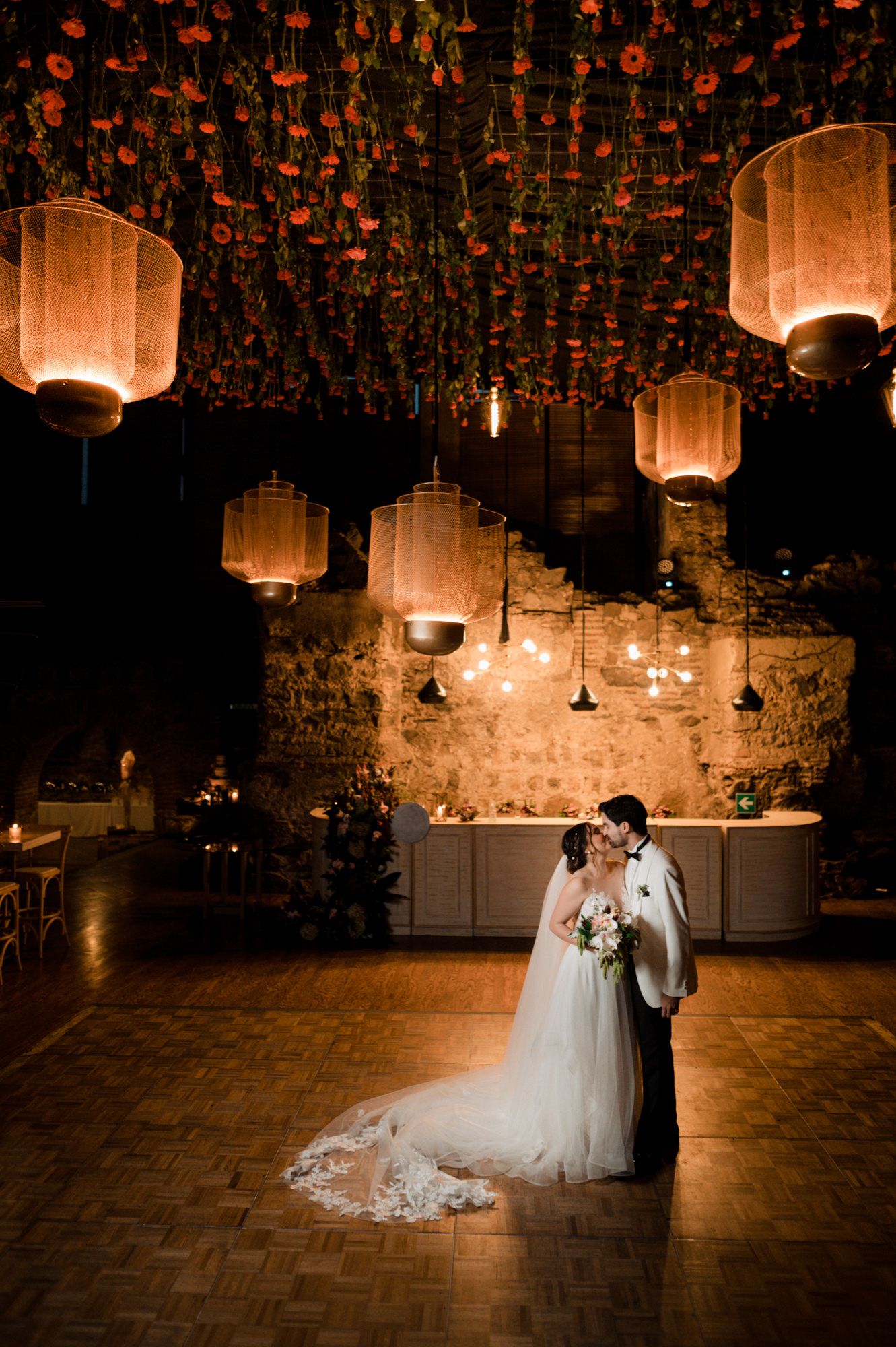 A romantic wedding scene with glowing lanterns overhead and a couple dancing in an elegantly lit stone venue.