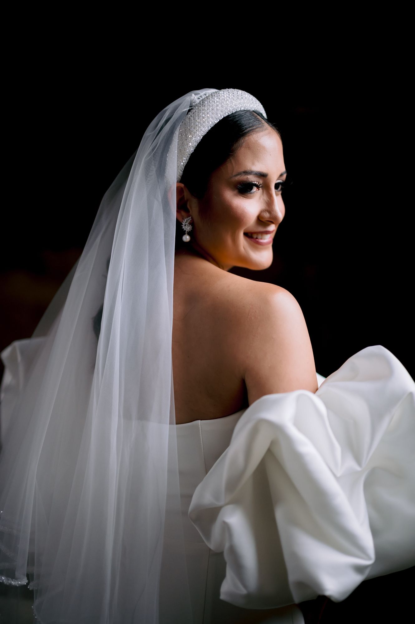 Bride in elegant white dress and veil smiles over shoulder in dramatic portrait lighting.