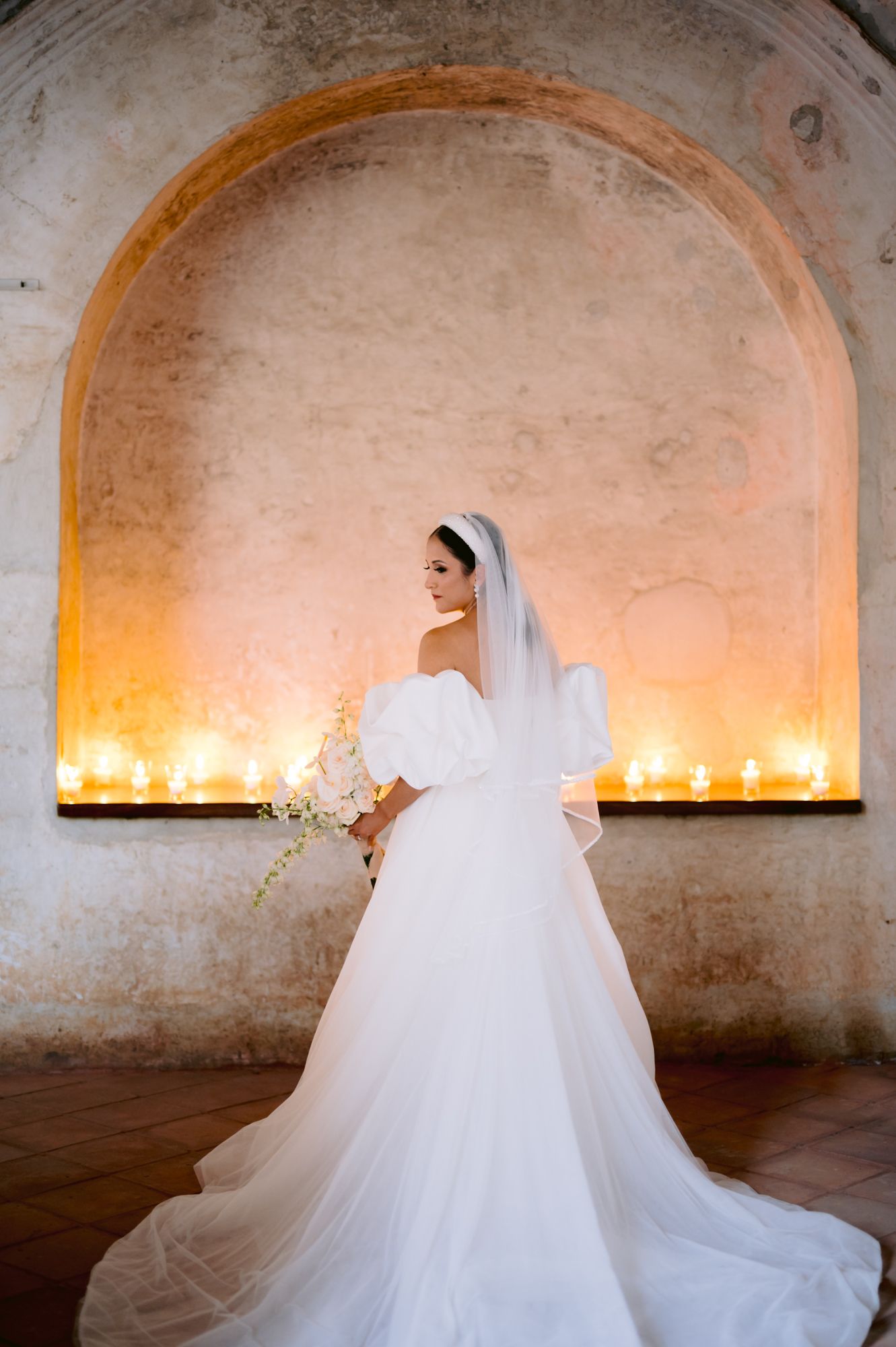 Elegant bridal portrait under candlelit stone arch showcasing off-shoulder gown with dramatic train.