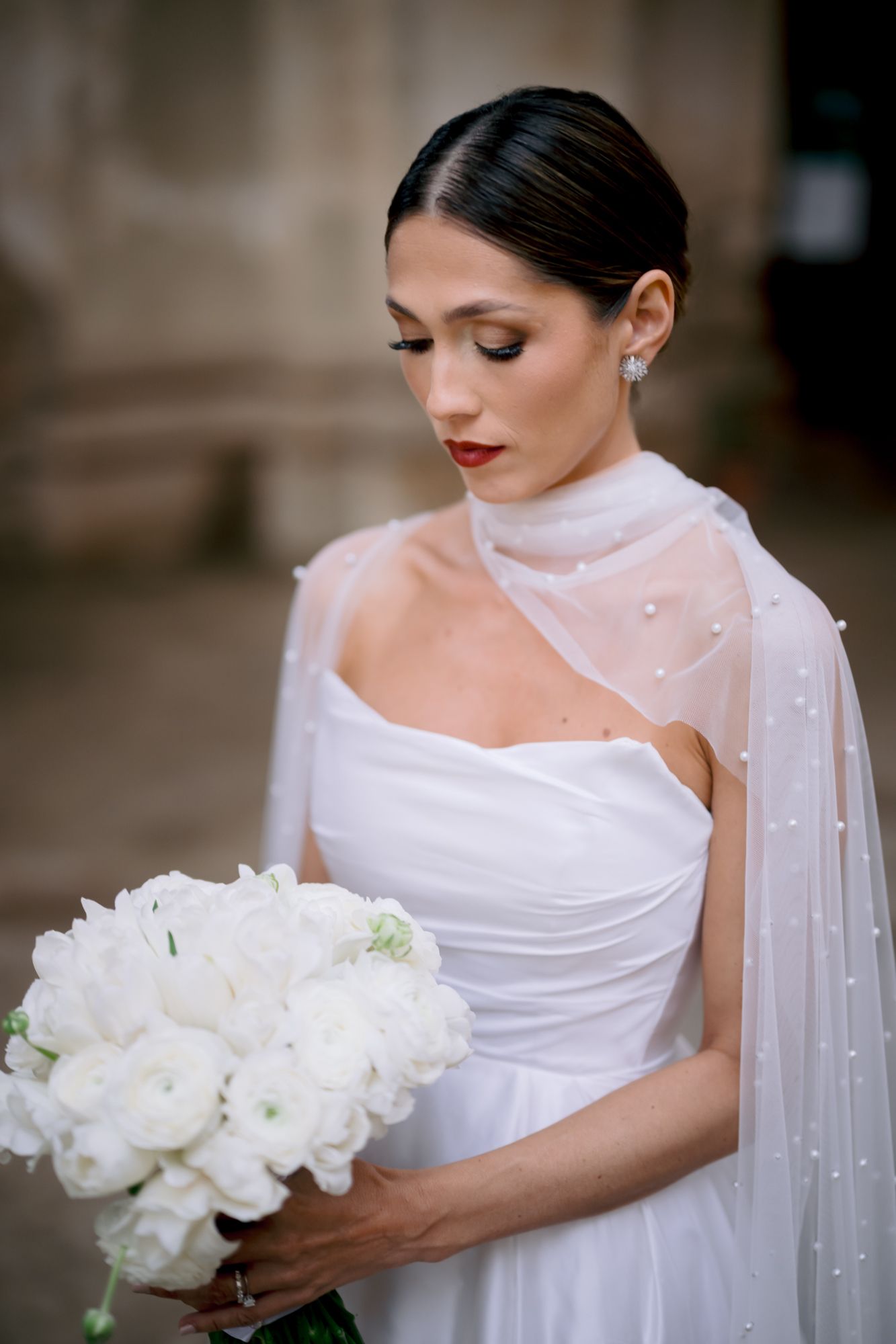 Elegant bride in white strapless gown holds luxurious white bouquet while looking down.