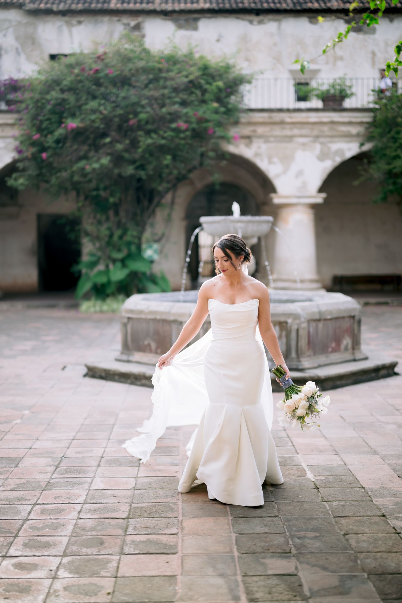 A bride walks gracefully through a courtyard with arches and fountain, holding her bouquet.