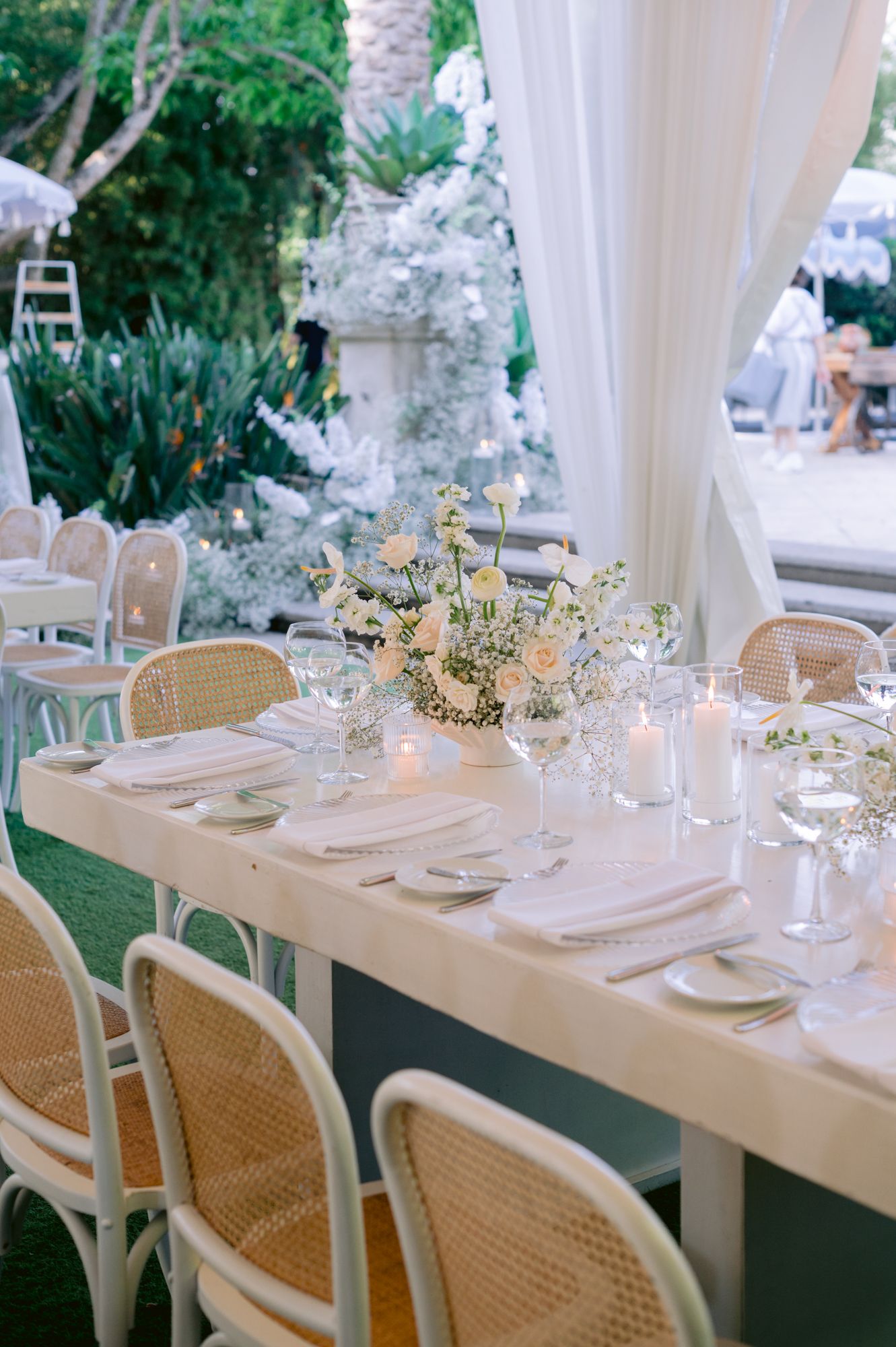 Elegant outdoor wedding table setting with white linens, cane chairs, and delicate floral centerpieces.