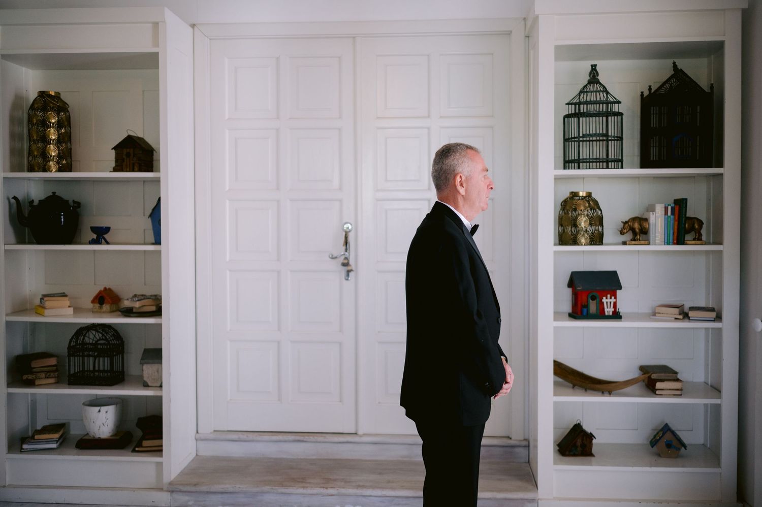 Man in black suit standing in profile against white built-in shelving.