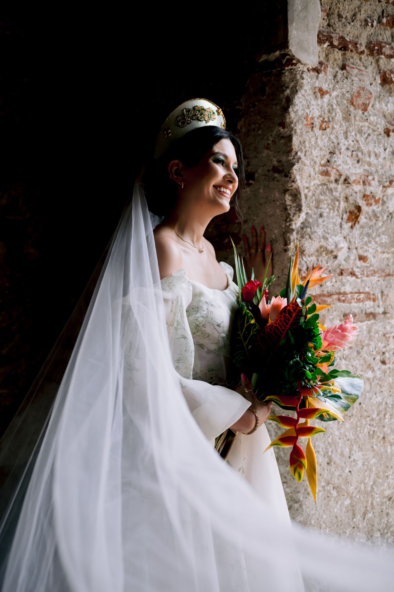 Bride wearing decorative crown holding vibrant tropical bouquet against rustic wall.