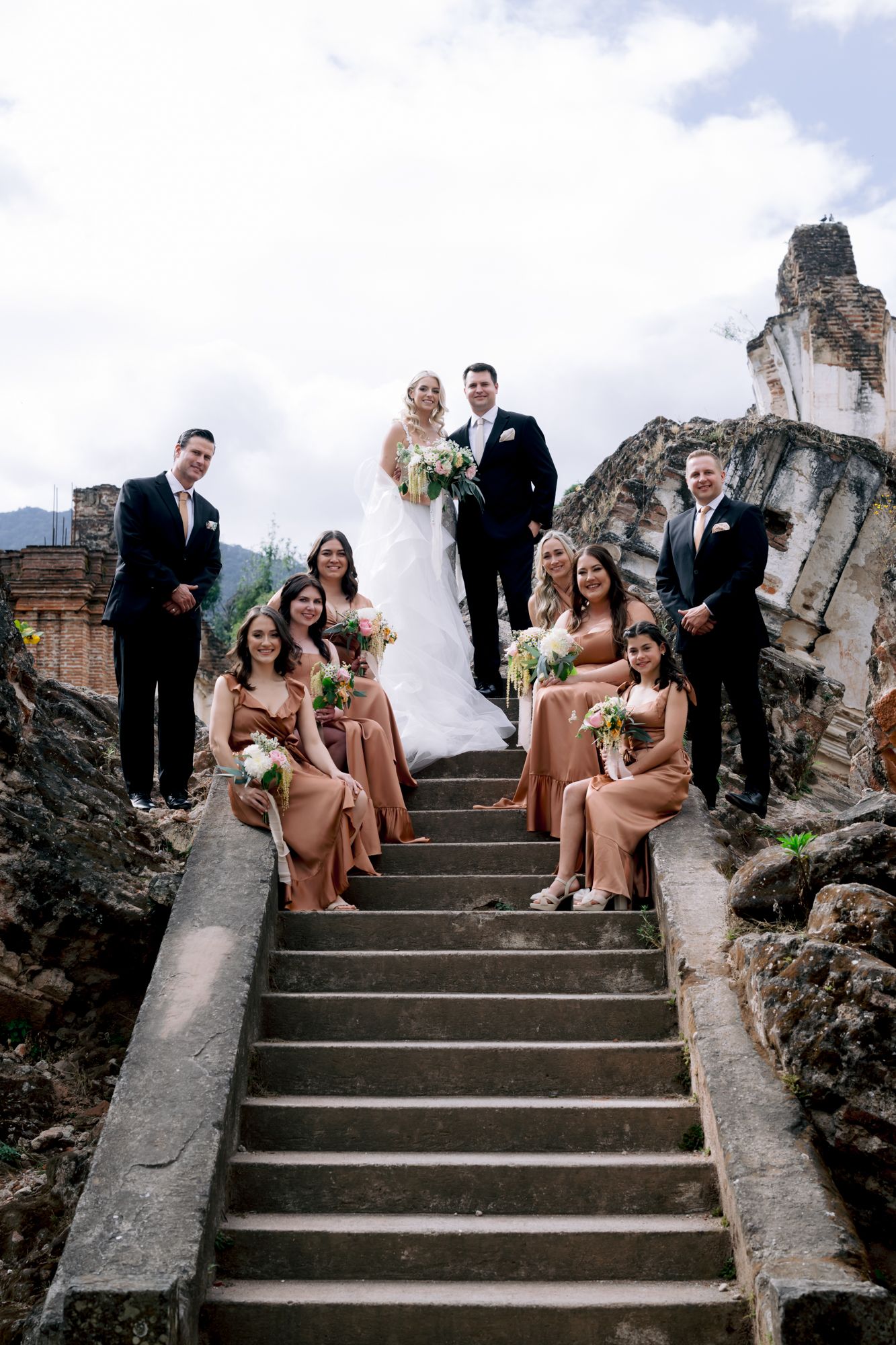Wedding party poses on stone steps with ruins and mountains in background.