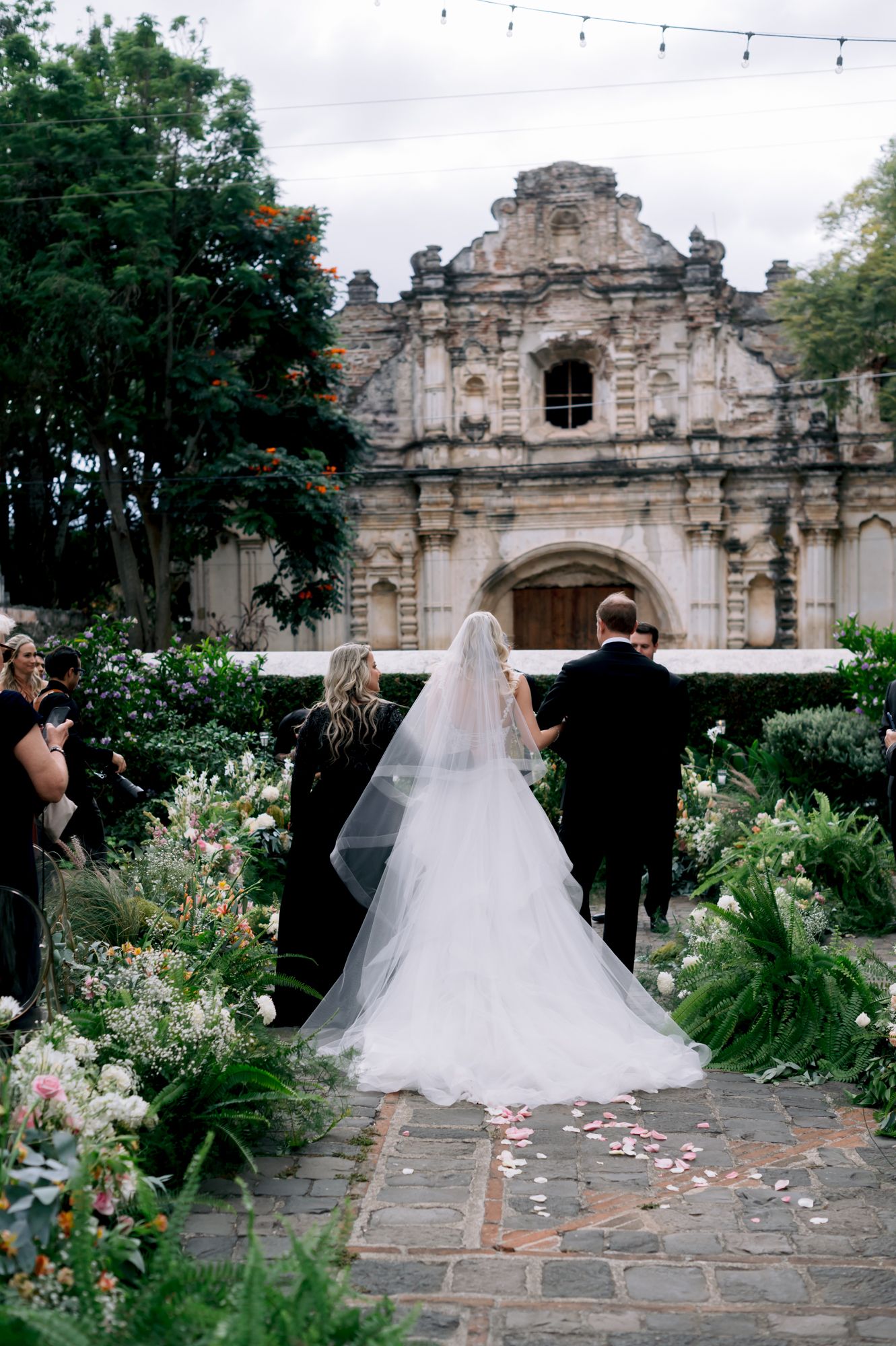 Wedding ceremony setup with flowing white dress against backdrop of historic stone church and garden decor.