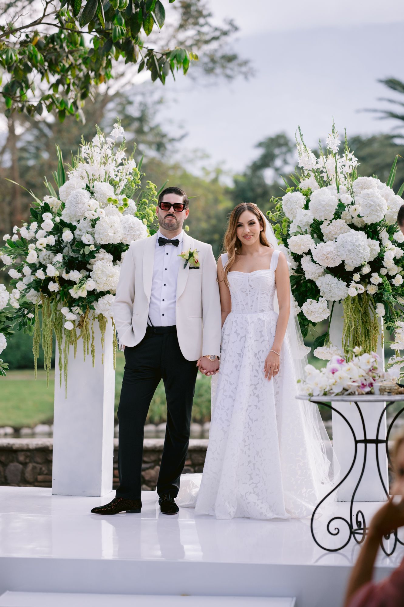 Elegant wedding ceremony setup with white floral arrangements and pedestals on a raised white platform outdoors.