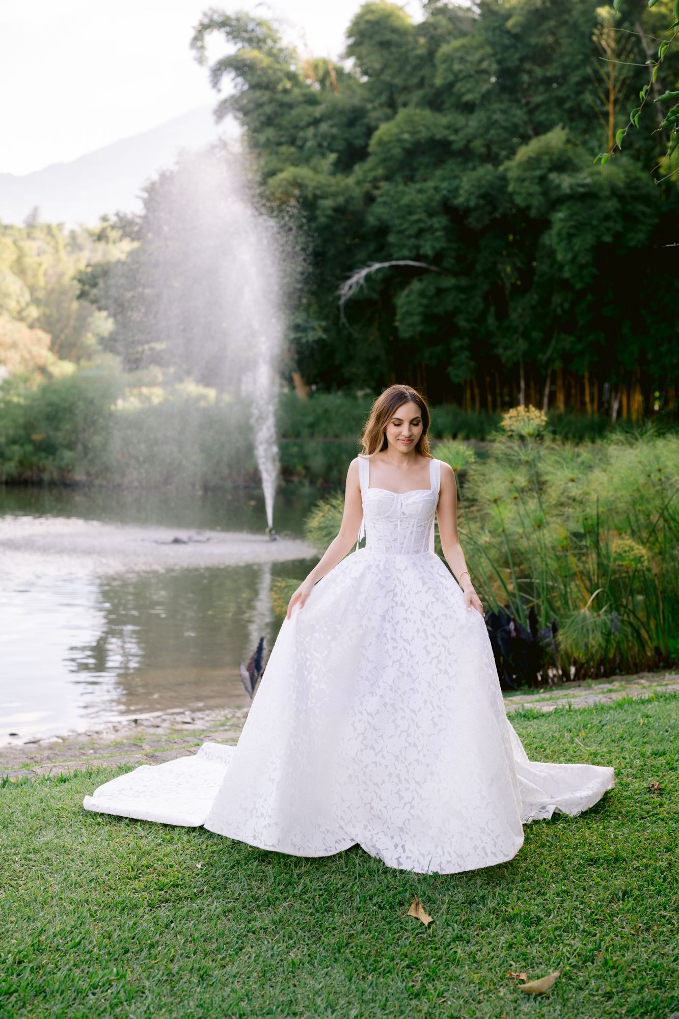A white wedding dress billows gracefully on a grassy lawn beside a tranquil pond with fountain spray in the background.
