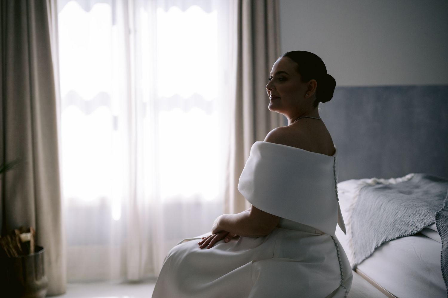 Silhouette by window in white off-shoulder dress during quiet morning preparation.
