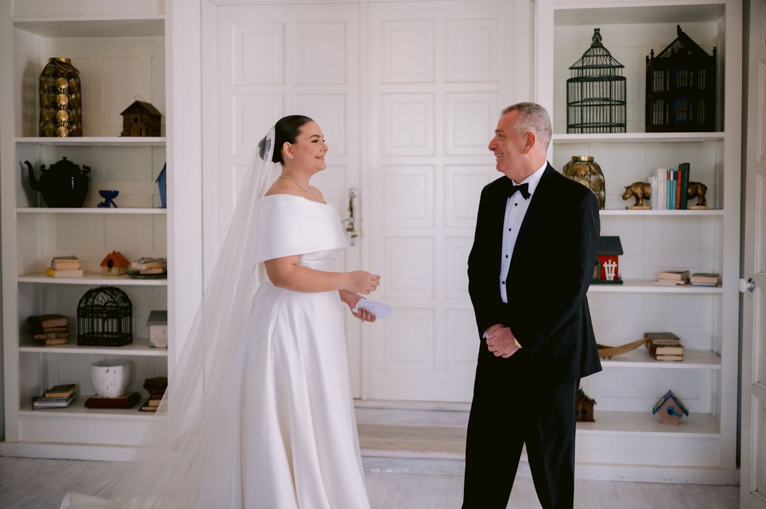 A bride in a white dress shares a lighthearted moment with a gentleman in a tuxedo against a white wall with shelves.
