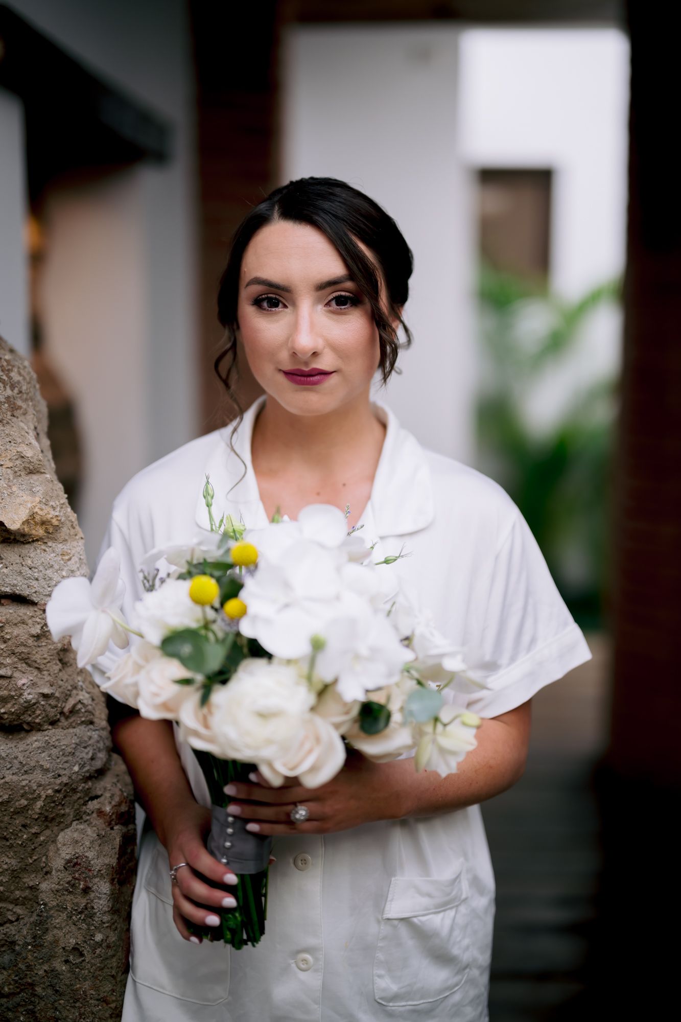 A person in a white robe holds a bouquet of white flowers against a rustic stone wall.