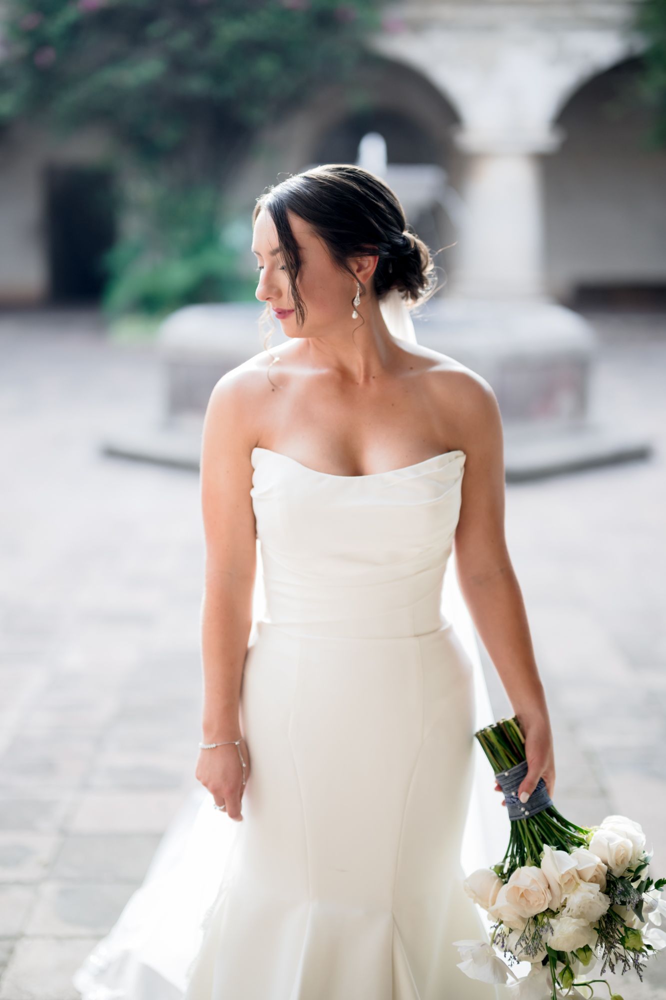 A radiant bride in a strapless gown holds a white bouquet in a historic courtyard.