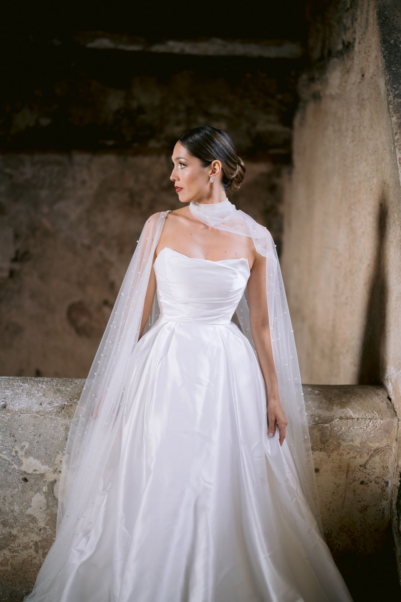 An elegant white wedding gown with a flowing tulle cape photographed against a rustic stone wall.