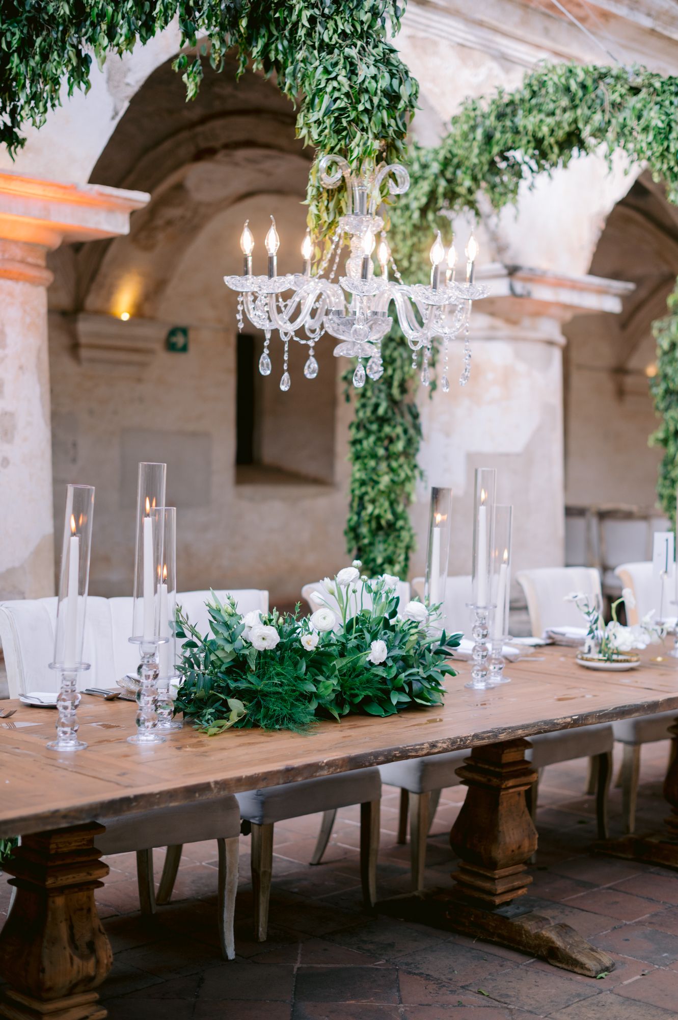 Elegant table setting with crystal chandelier and greenery centerpiece in rustic courtyard.