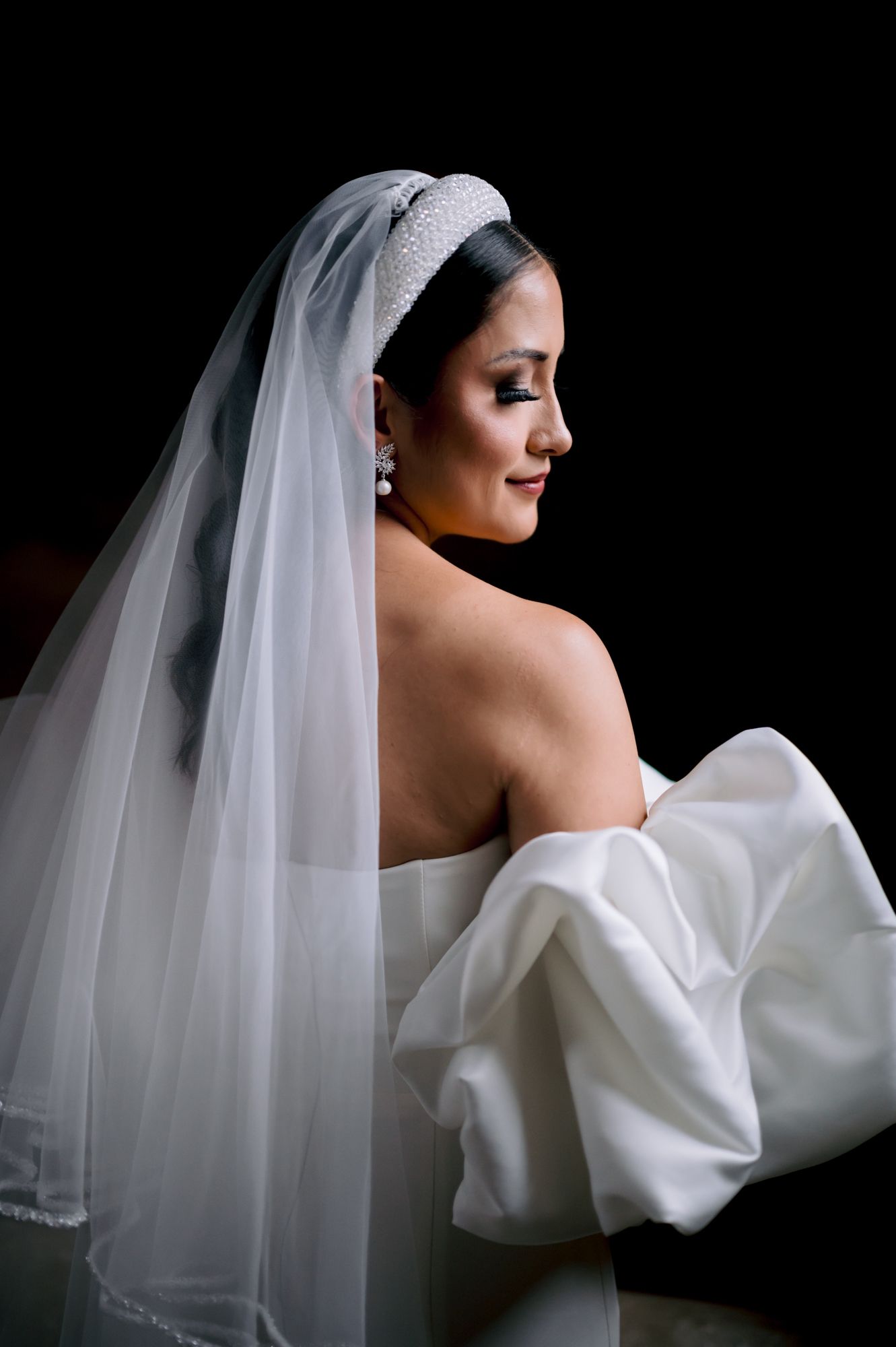 A dramatic side profile portrait of a bride wearing an off-shoulder gown and flowing veil against black background.