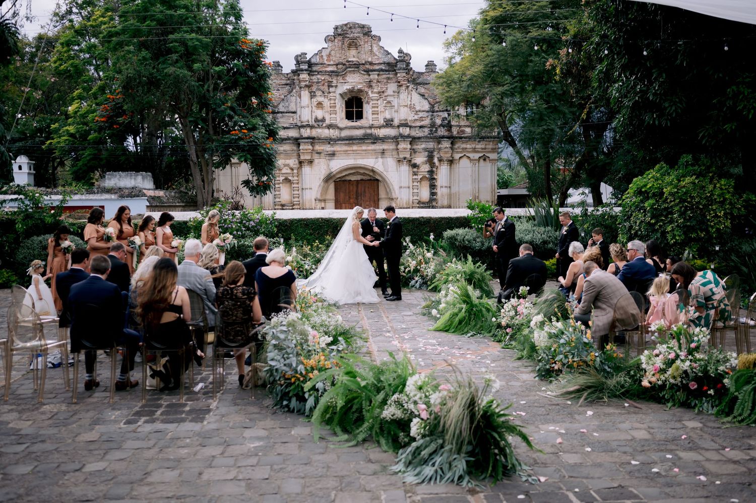 Wedding guests seated outdoors along flower-lined aisle with historic stone church as backdrop.