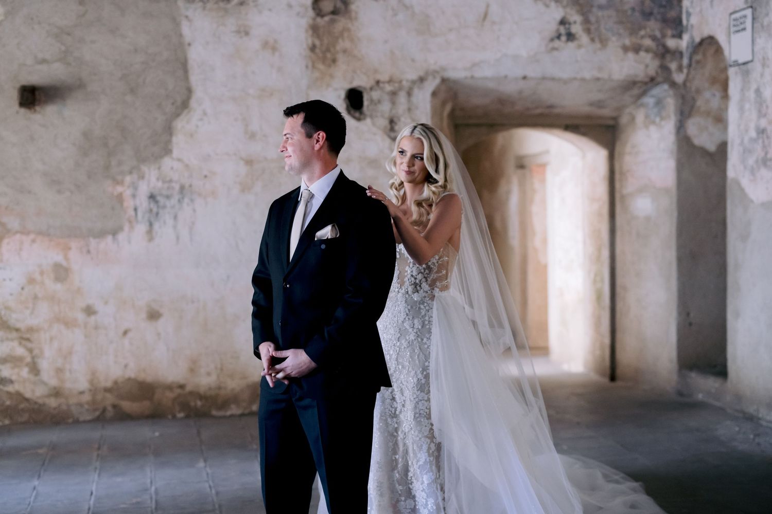 Bride playfully peaks at groom during first look in rustic stone venue.