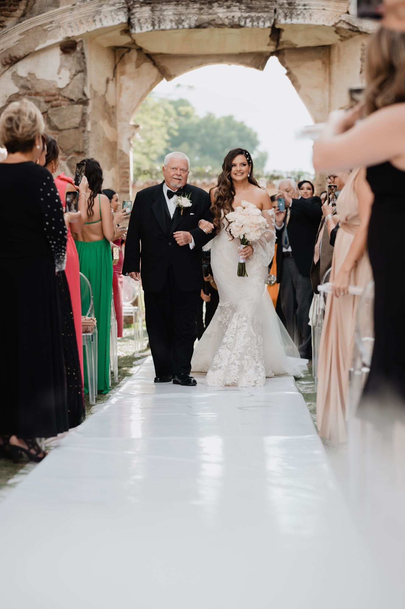 A bride in a mermaid wedding gown walks down a white aisle with guests in formal attire watching from both sides.
