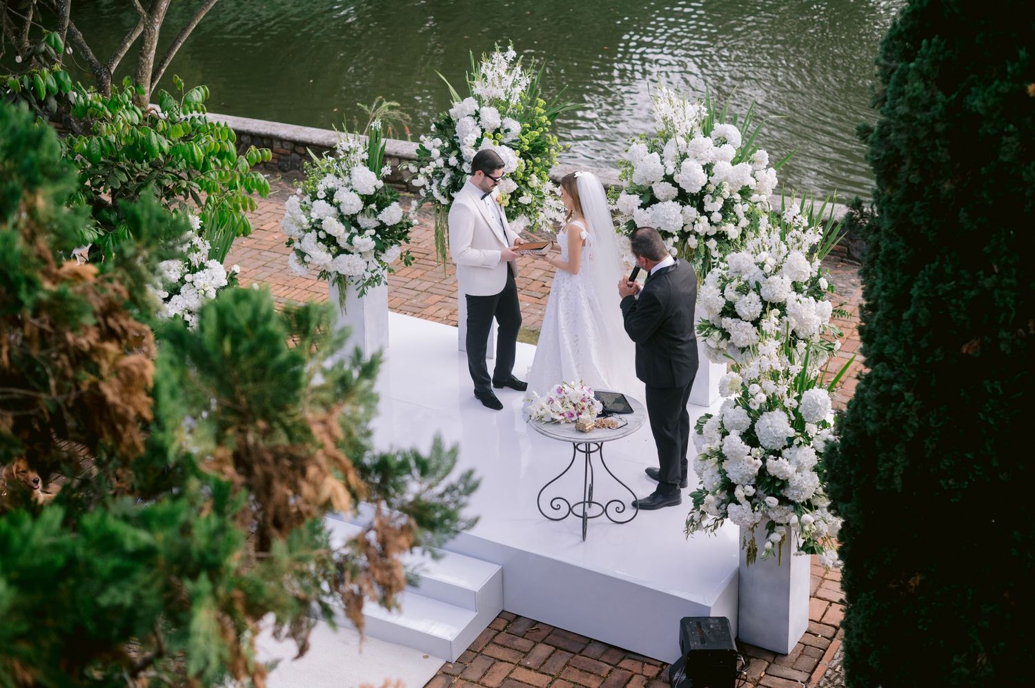 An outdoor wedding ceremony setup with a white floral arch overlooking a lake, featuring elegant white decorations.