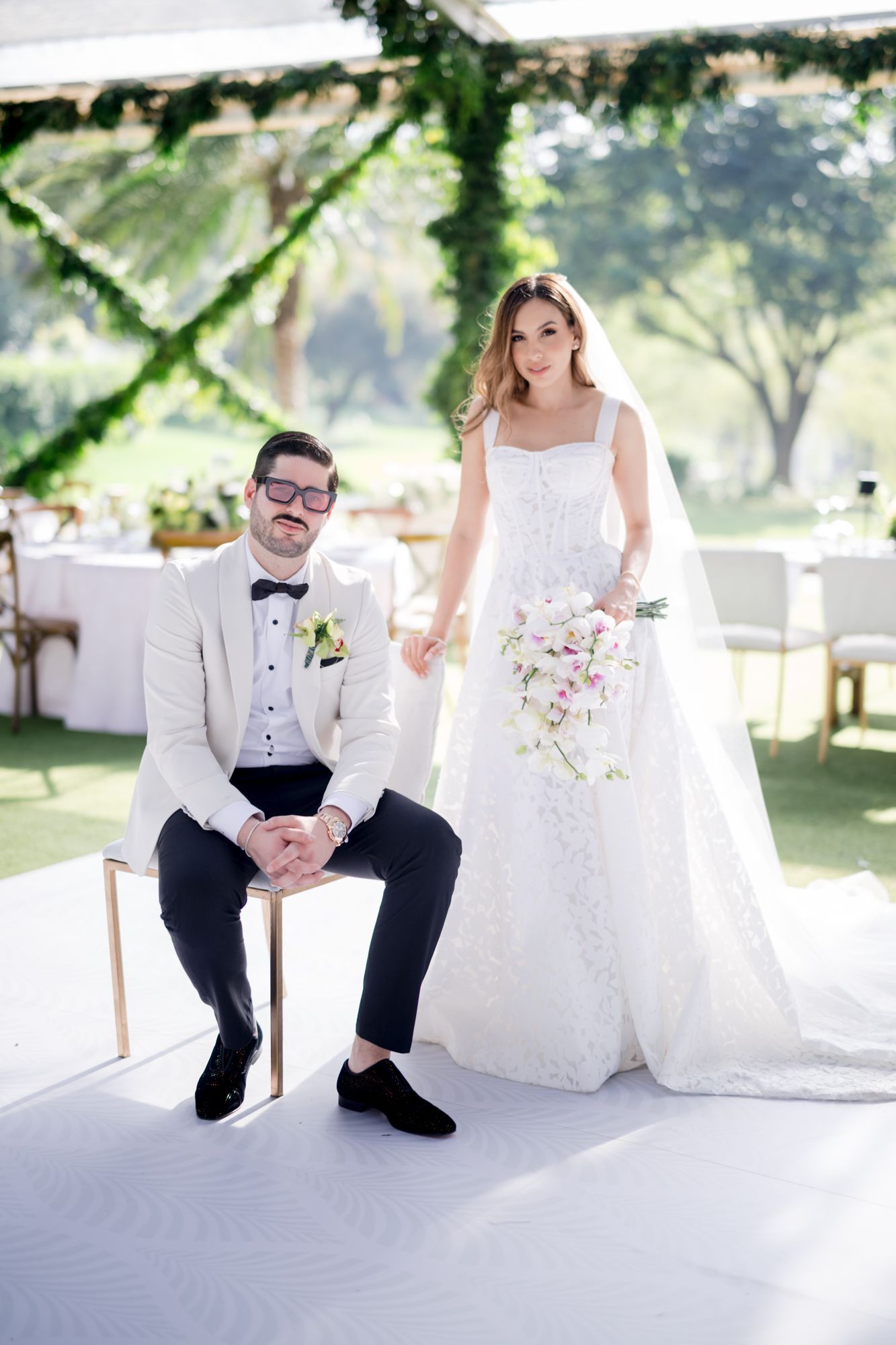 Elegant outdoor wedding portrait with couple in white attire standing on white platform with floral decor.