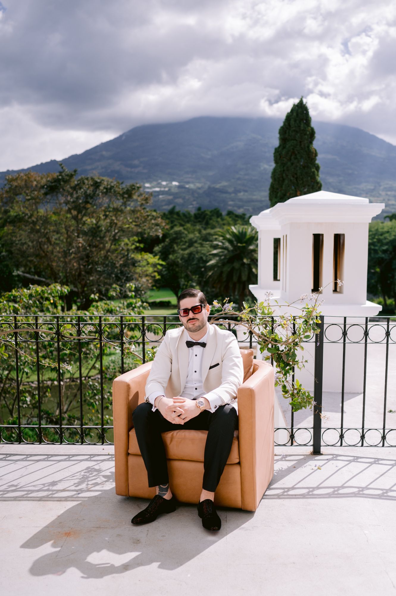 A person in formal attire sits casually on a tan leather chair on a white terrace with mountain views in the background.