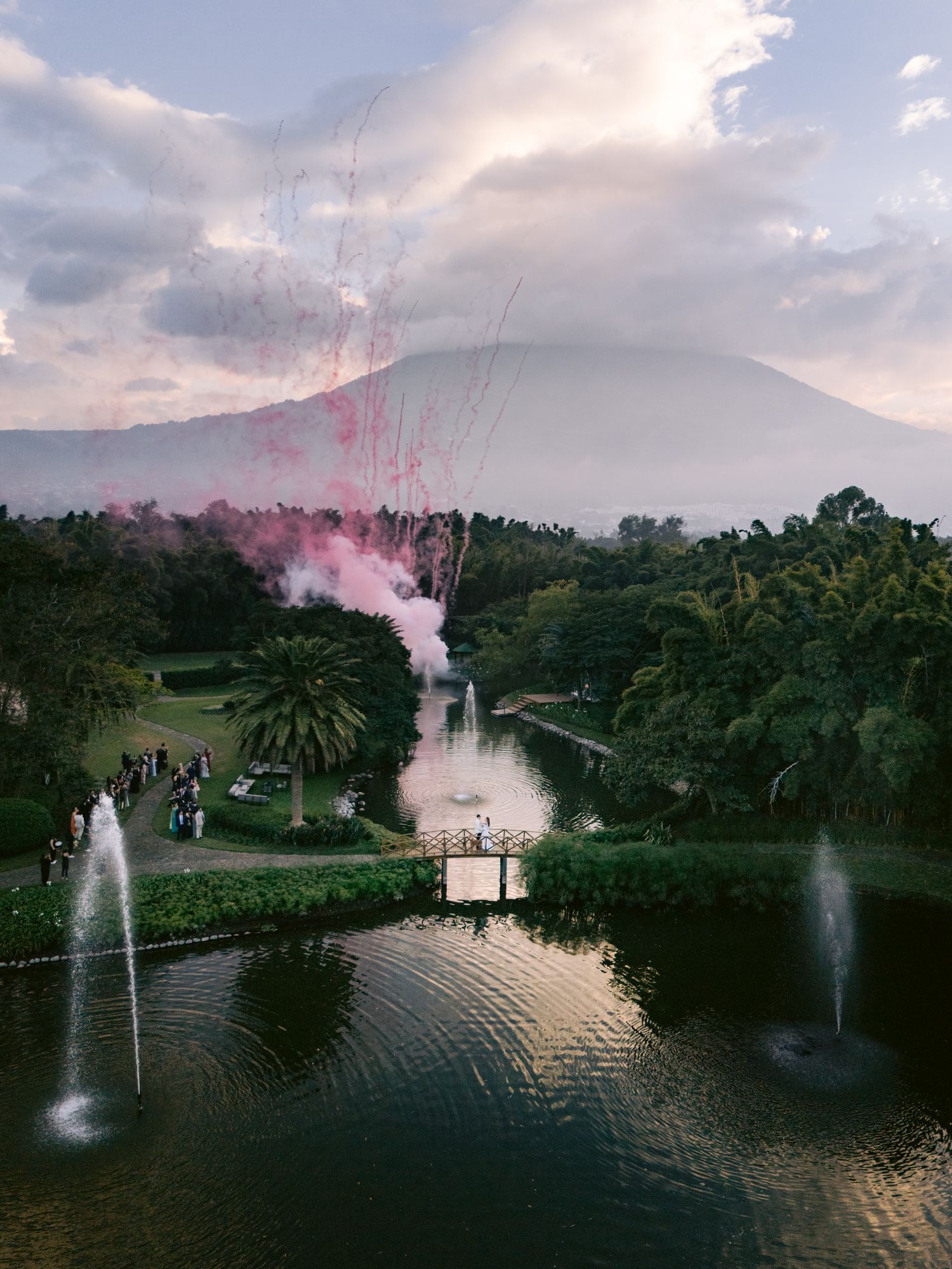 Scenic garden pond with fountains and pink smoke against a mountain backdrop at sunset.