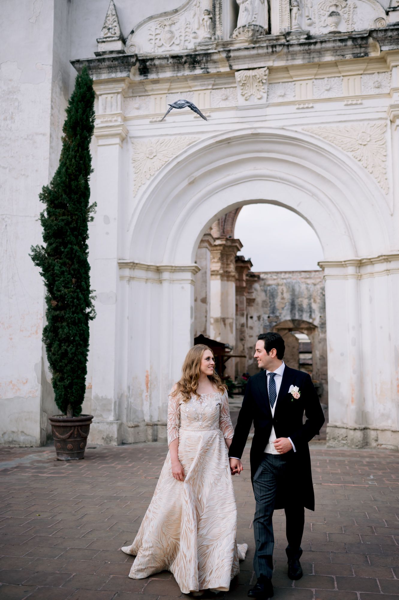Couple walking together outside ornate white archway with cypress tree.