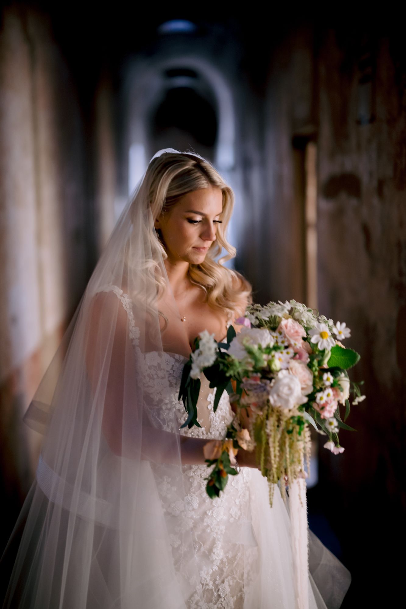 An elegant bridal portrait showcasing a stunning lace wedding gown and delicate floral bouquet in a dramatic hallway.