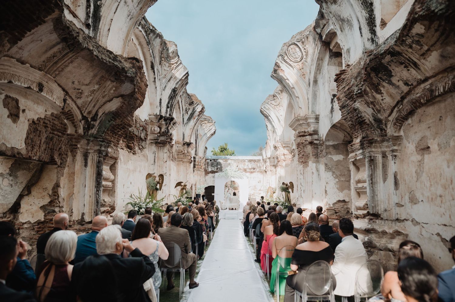 A wedding ceremony takes place in a historic ruins with stone archways and exposed sky overhead as guests watch from wooden chairs.