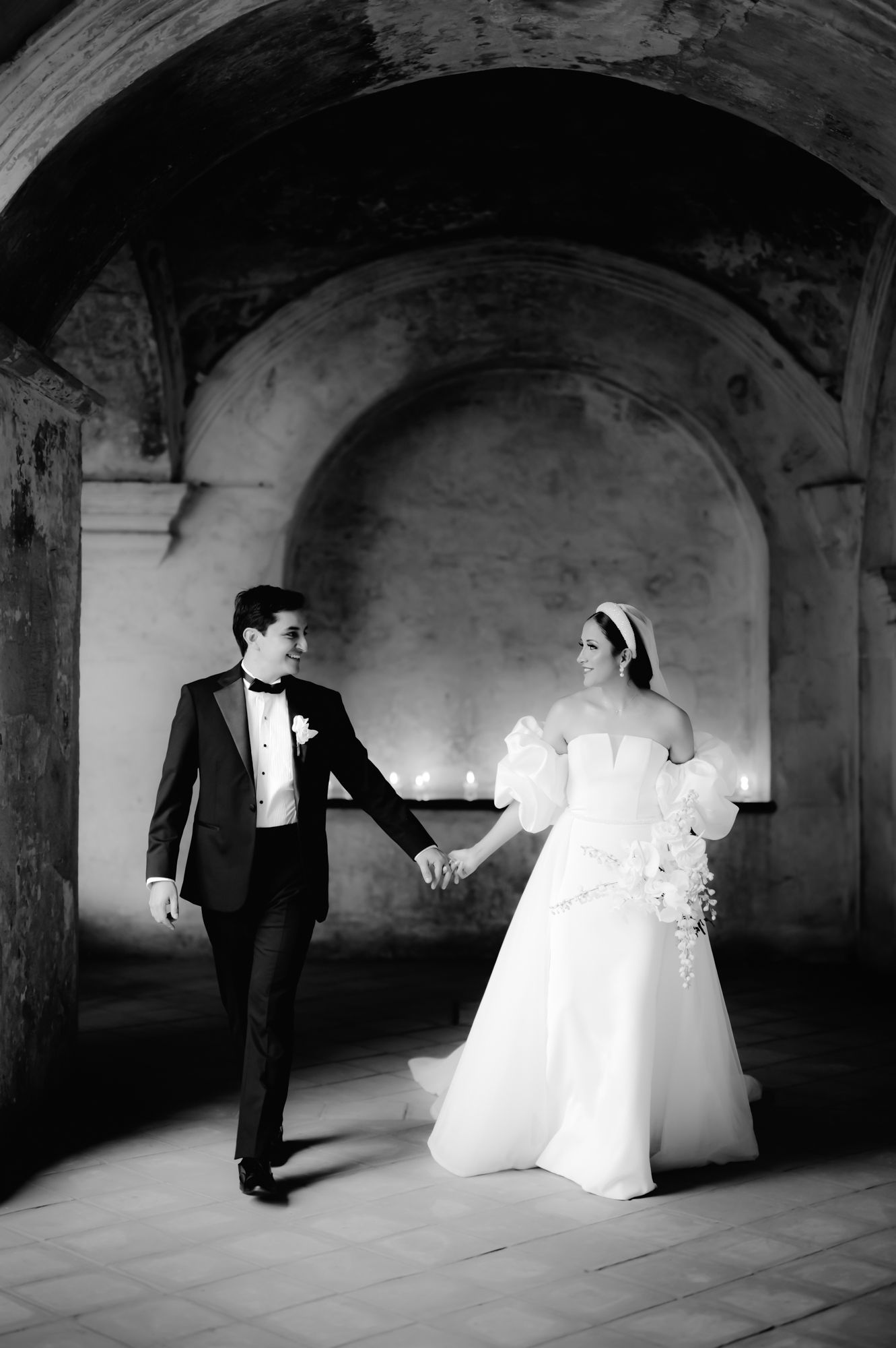 A black and white photo of newlyweds holding hands while walking under a historic arched corridor.