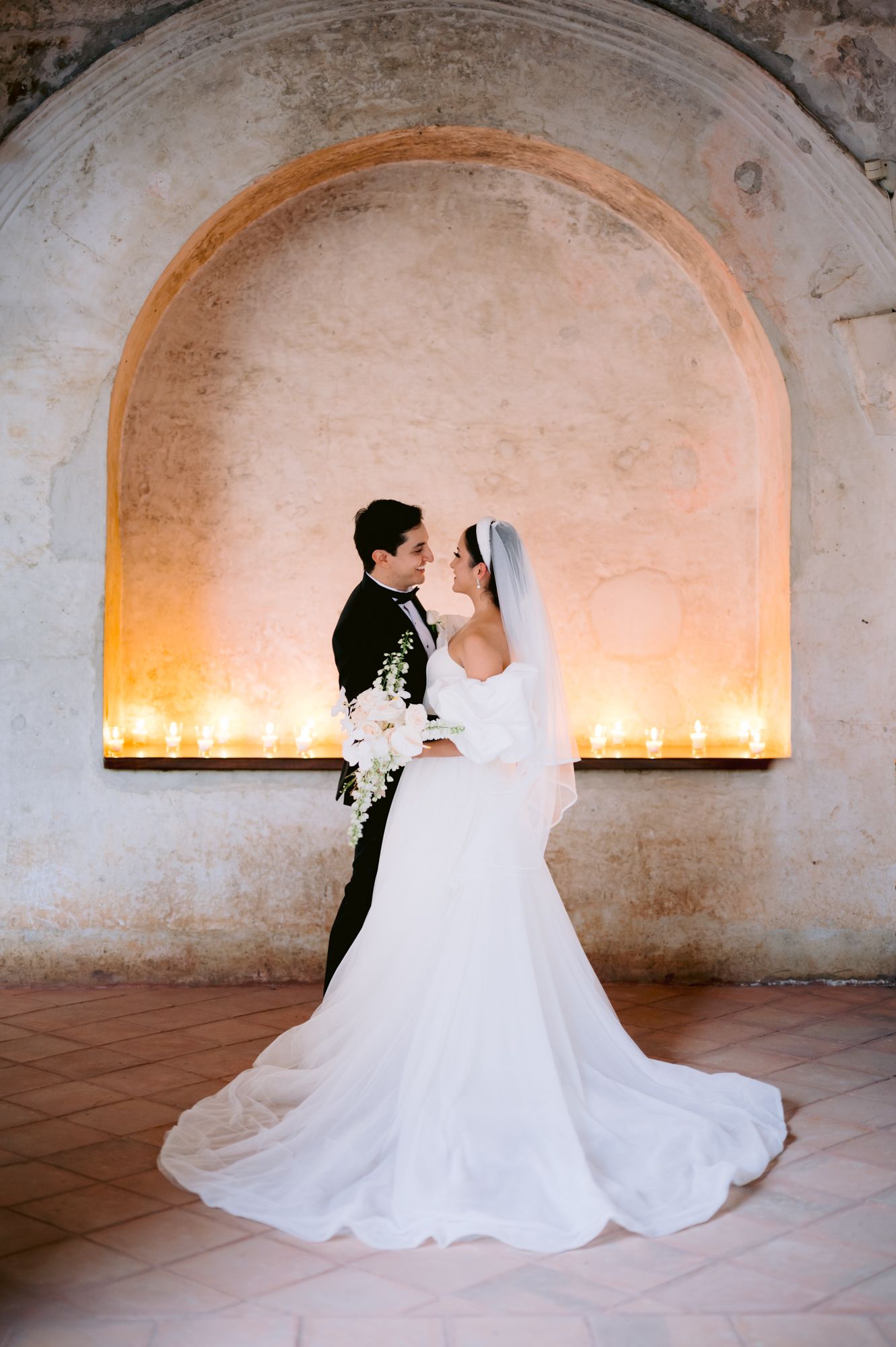 Romantic wedding couple poses in candlelit stone archway during evening ceremony.