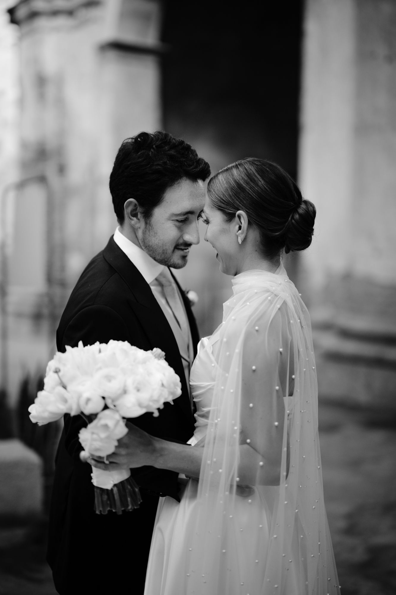 A romantic wedding moment with the bride in a delicate veil holding white flowers while standing close to her groom.