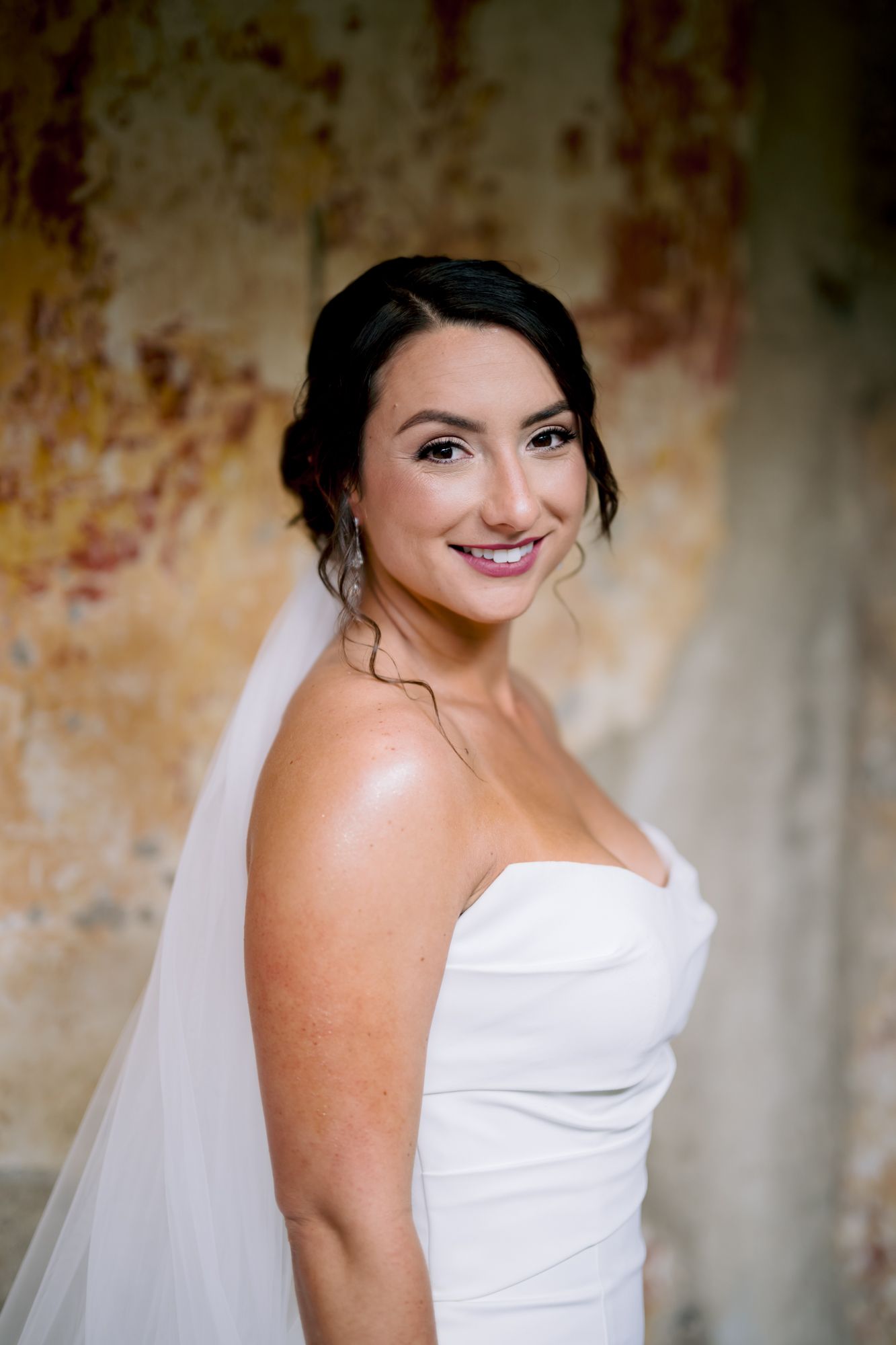A bride in a strapless white gown smiles in front of a distressed wall.
