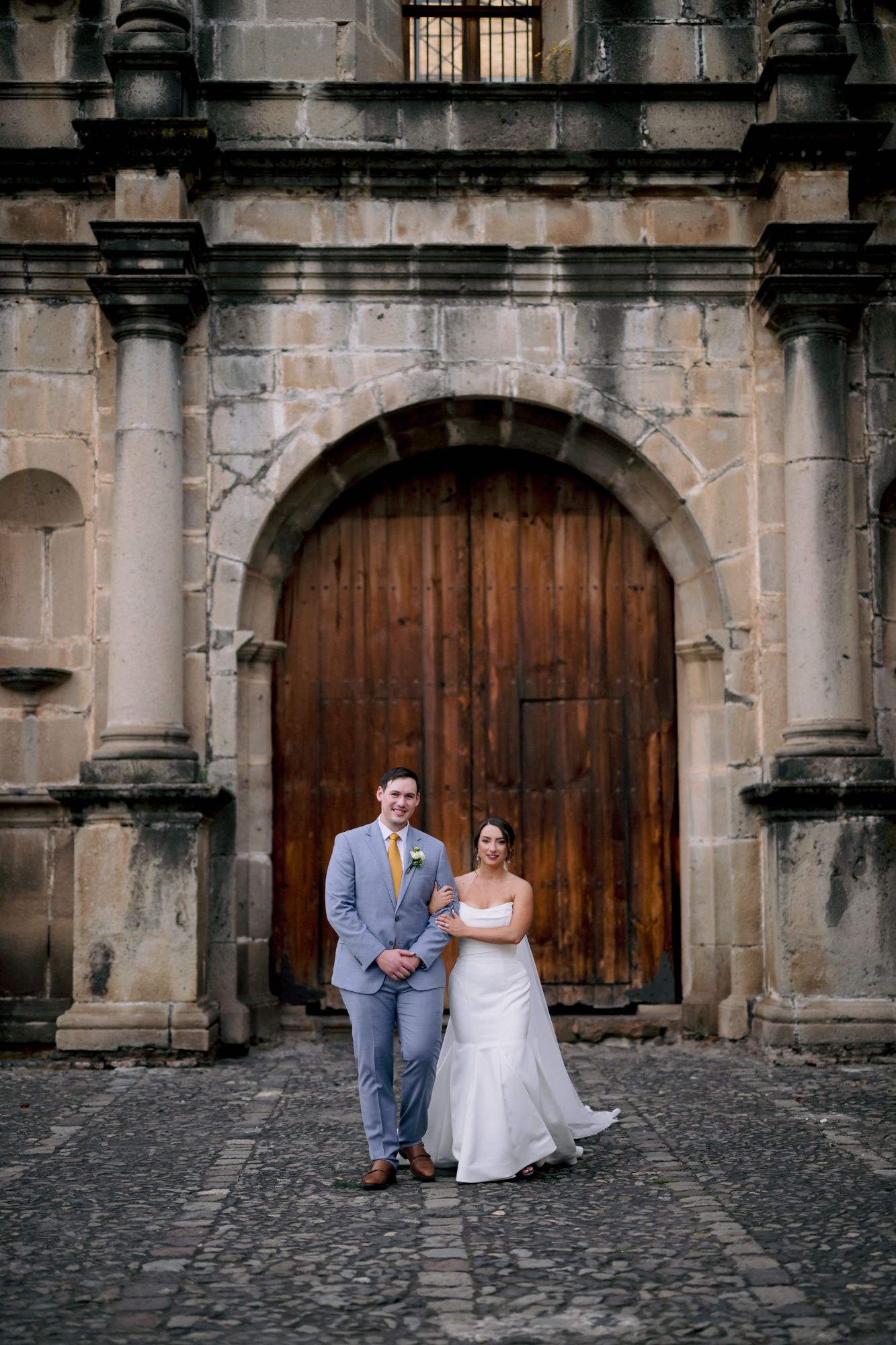 A wedding couple poses in front of a grand wooden door with ornate stone architecture.