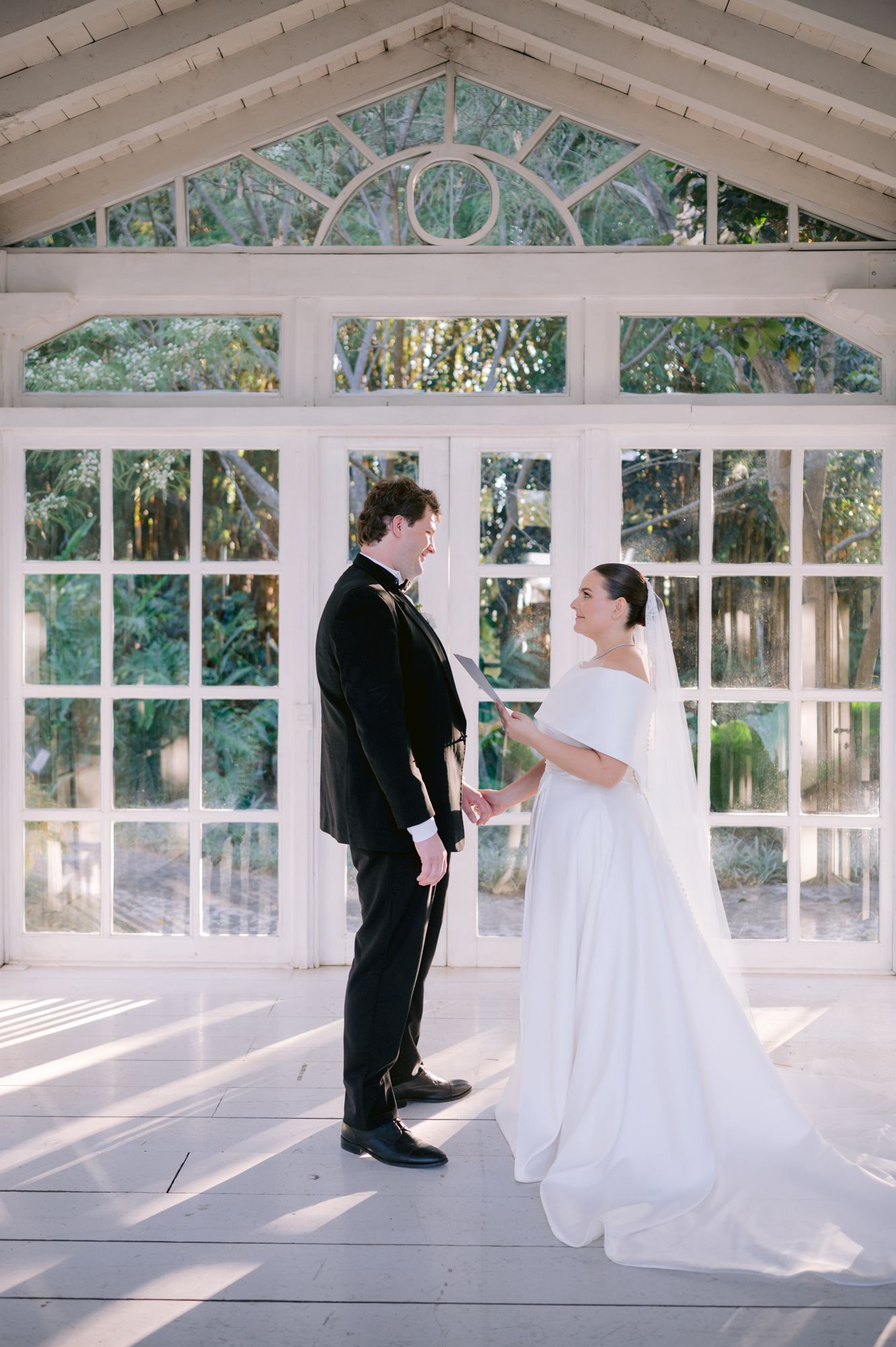 A wedding ceremony in a sunlit greenhouse with white architectural details and large windows.