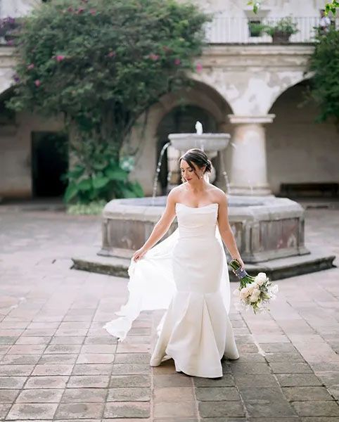 A bride in a flowing white wedding dress stands in a historic courtyard with stone arches and greenery.