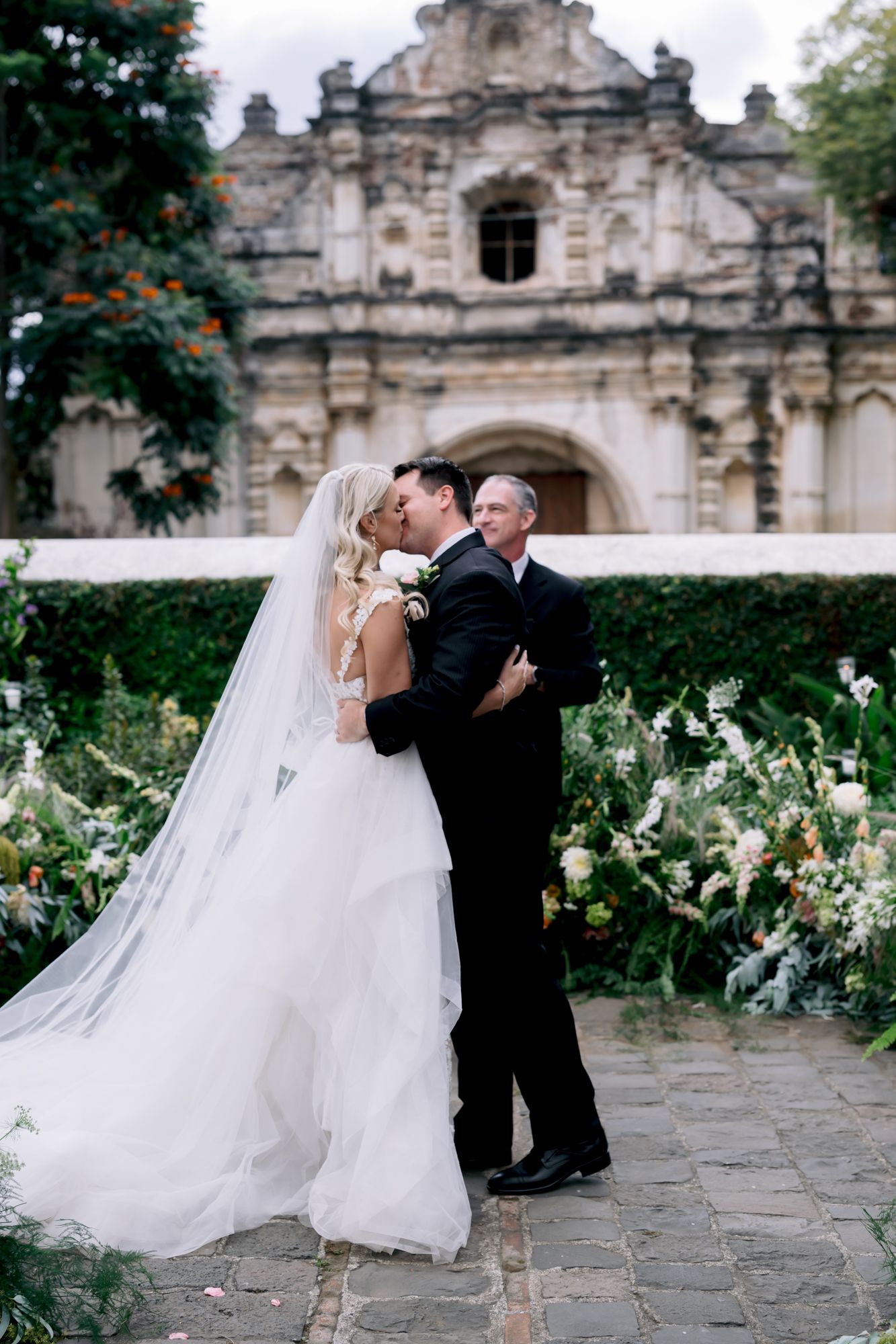 Romantic wedding moment captured in front of historic stone church with flowing veil and floral gardens.