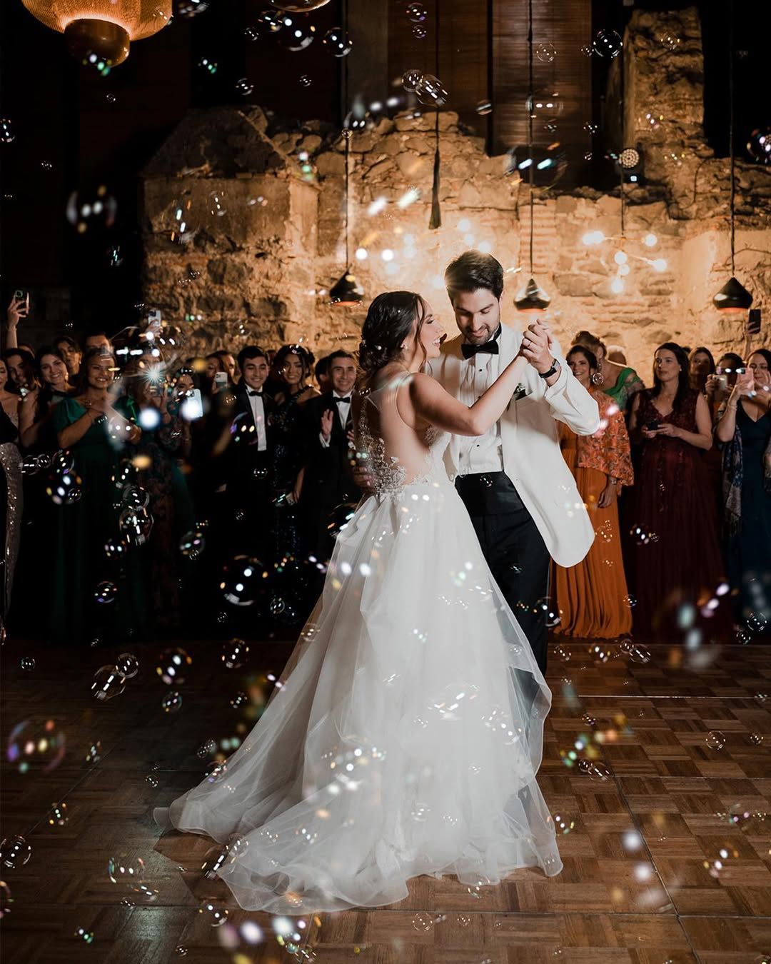 A wedding couple shares their first dance surrounded by floating bubbles in a romantic lit venue.