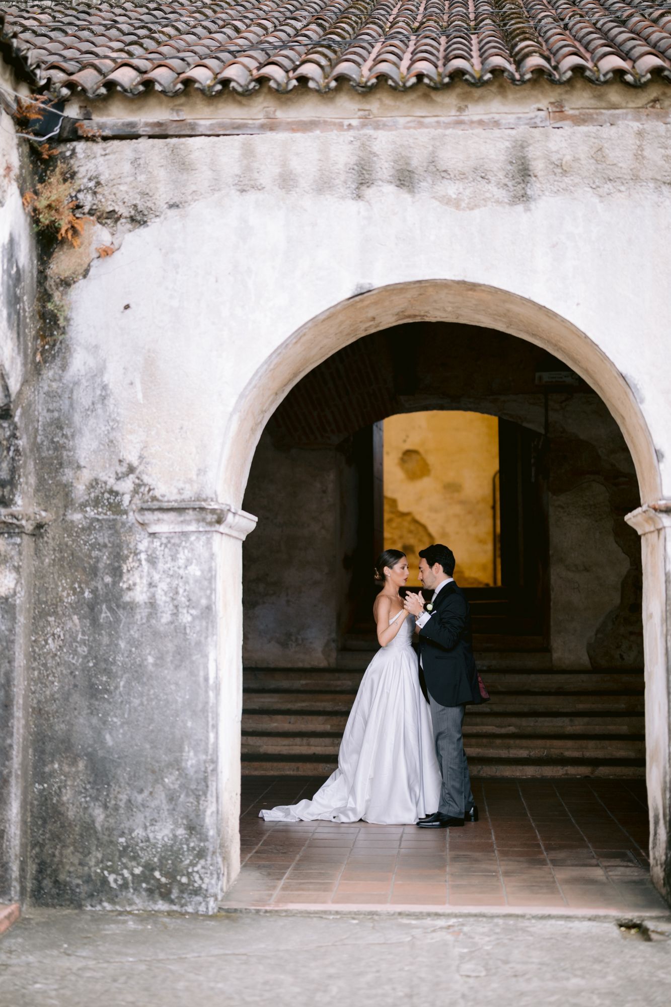 A couple shares an intimate moment under a rustic stone archway with terracotta tiles.