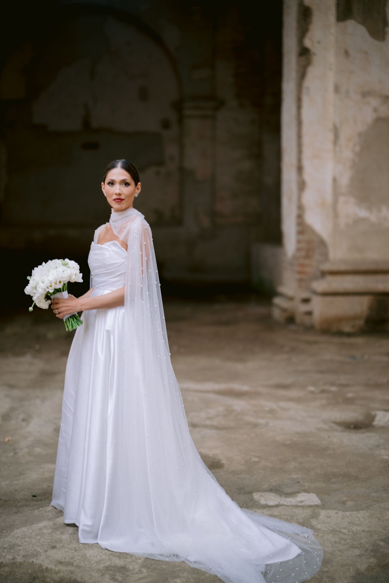 Bride in flowing white dress and cape holding white bouquet in historic stone building.