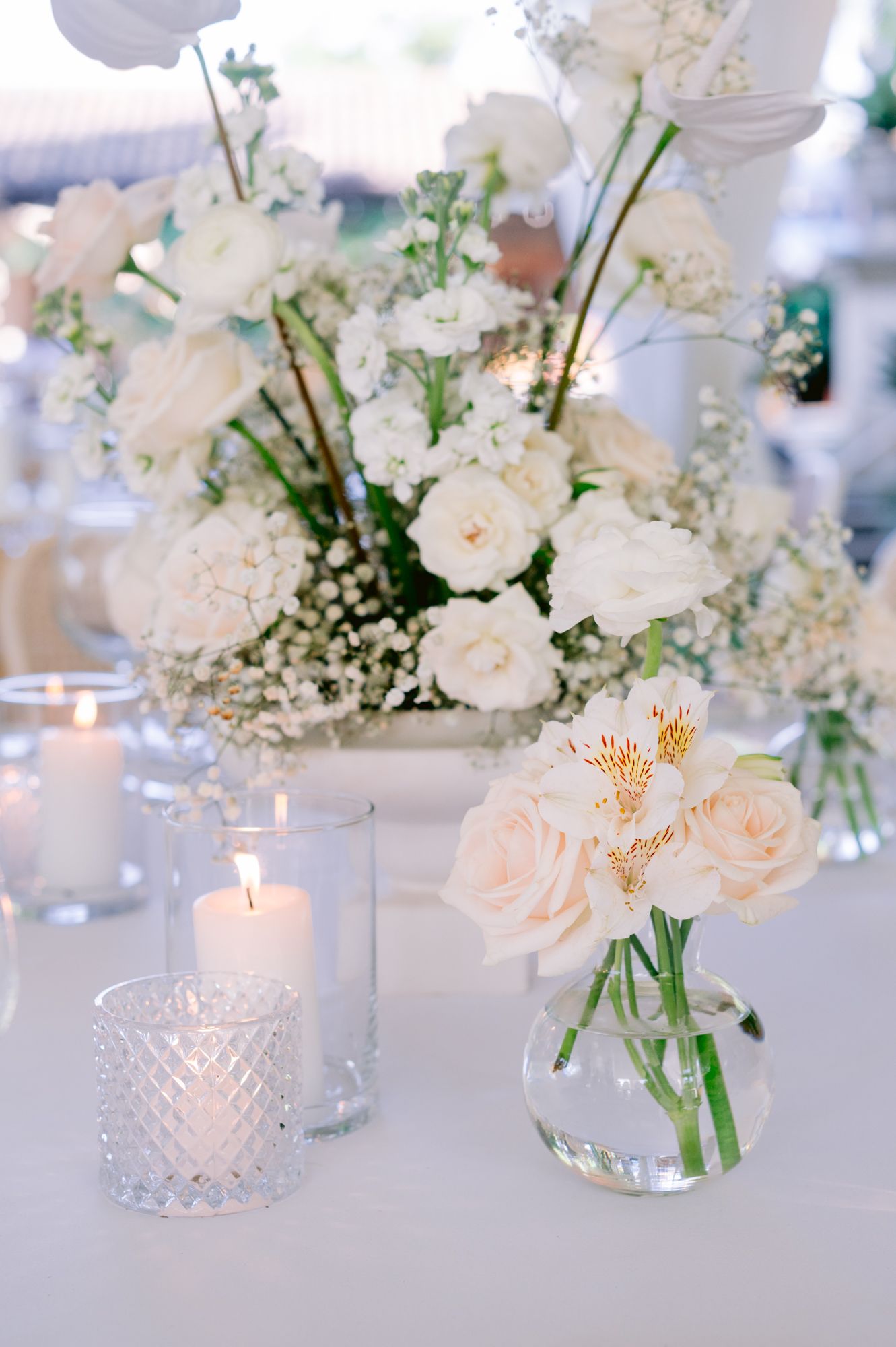 Romantic white floral centerpiece with roses and baby's breath alongside crystal candleholders on white tablecloth.
