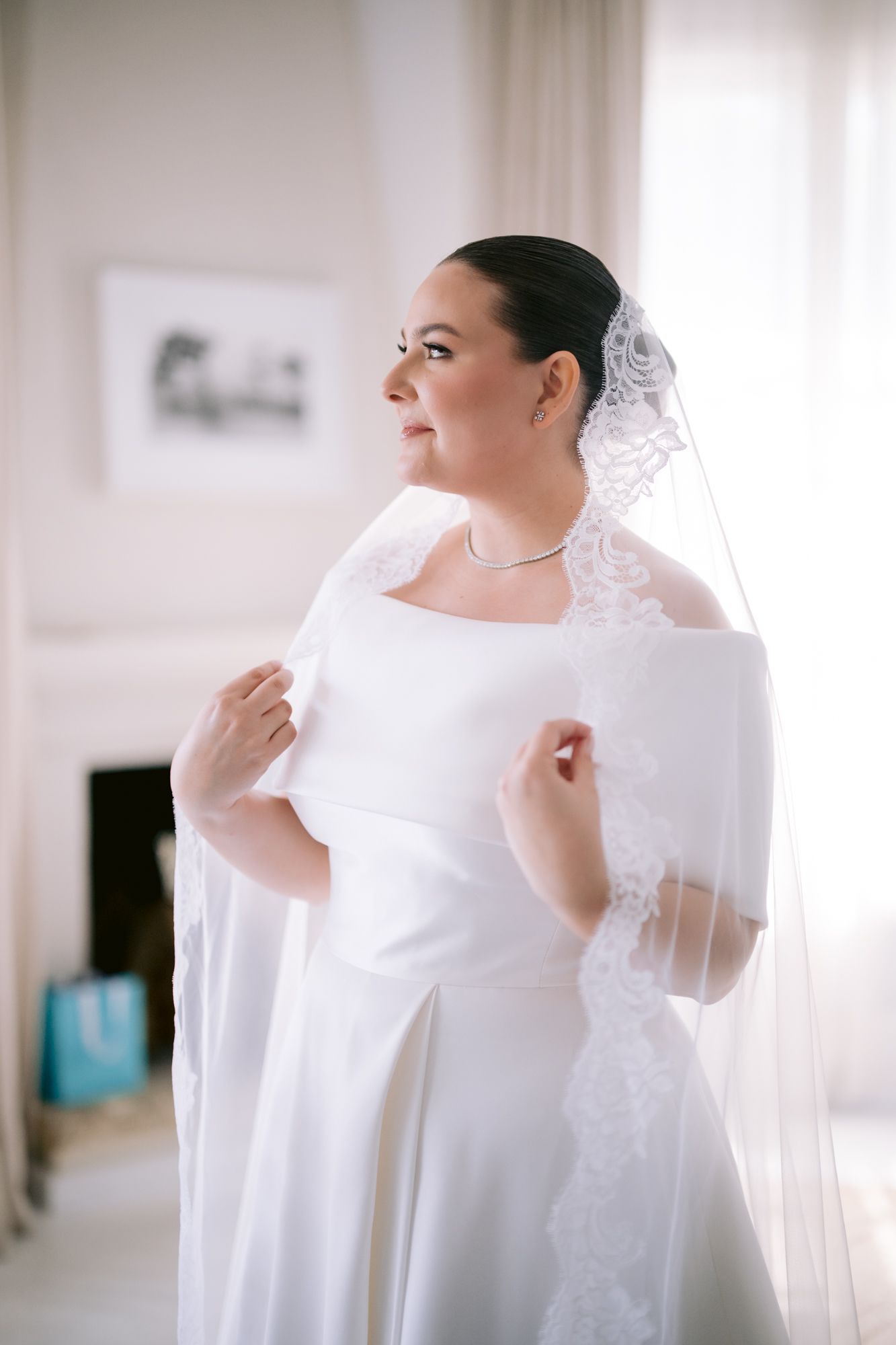 Bride in classic white off-shoulder gown with veil standing by window in bridal suite.