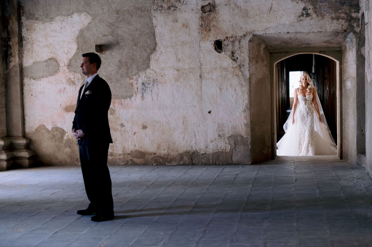 First look wedding photo with bride and groom standing apart in rustic stone hallway.