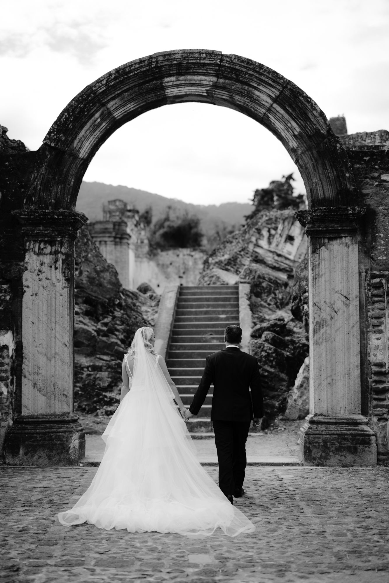 Black and white photo of newlyweds walking through stone archway toward grand staircase.