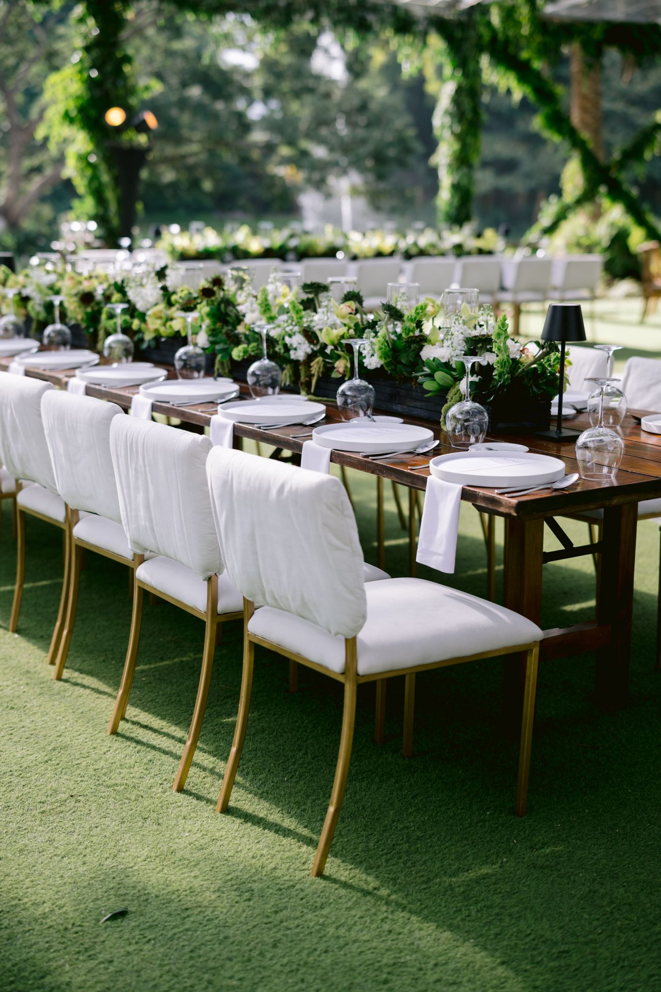 Elegant outdoor wedding reception setup with white cushioned chairs and wooden tables on green lawn under trees.