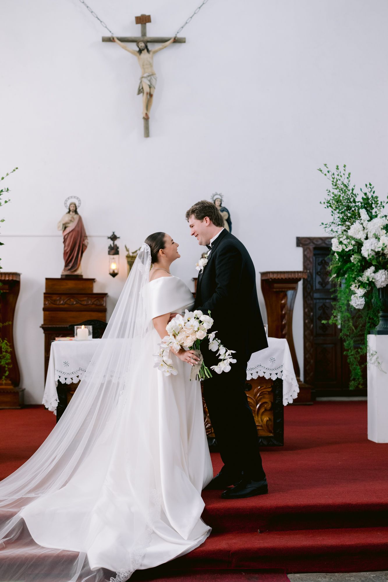 A romantic wedding moment in a traditional church with white floral arrangements and a crucifix above.